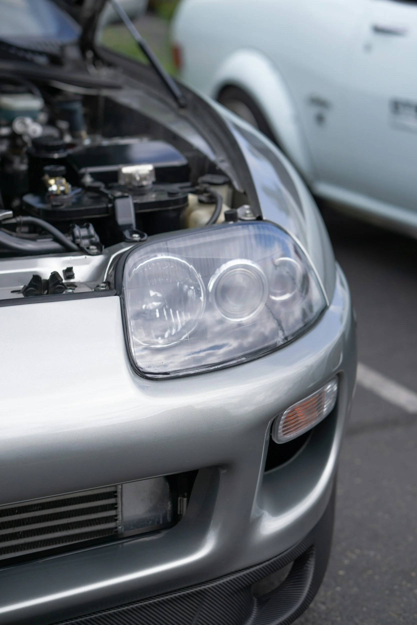 Close-up of a silver sports car with its hood open, showing the headlight and engine components, parked on a street.