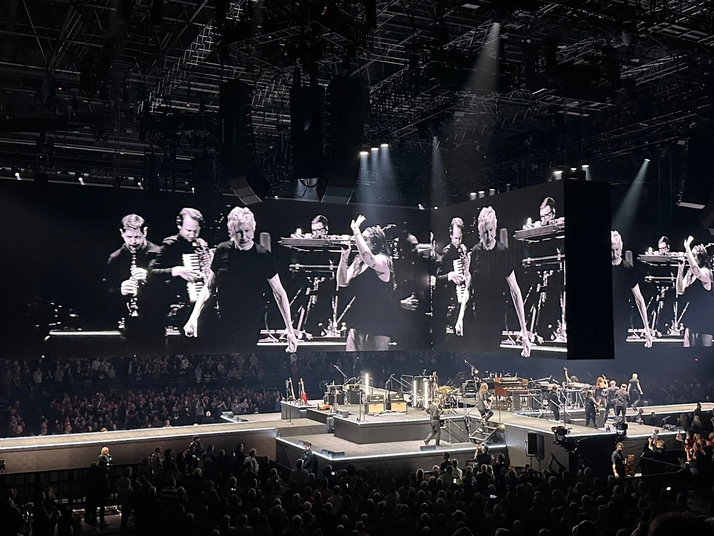 The touring band performing on the in-the-round stage with monochrome close-up footage projected across the overhead screens at Utilita Arena.