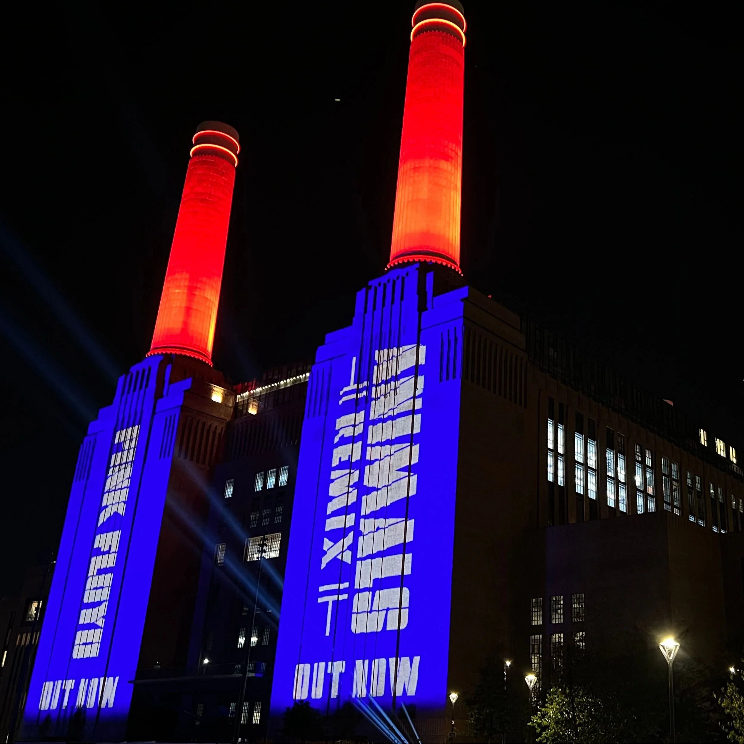 Close-up night shot of Battersea Power Station featuring bright red illuminated chimneys and blue projection mapping displaying 'Pink Floyd Animals Remix - Out Now'.