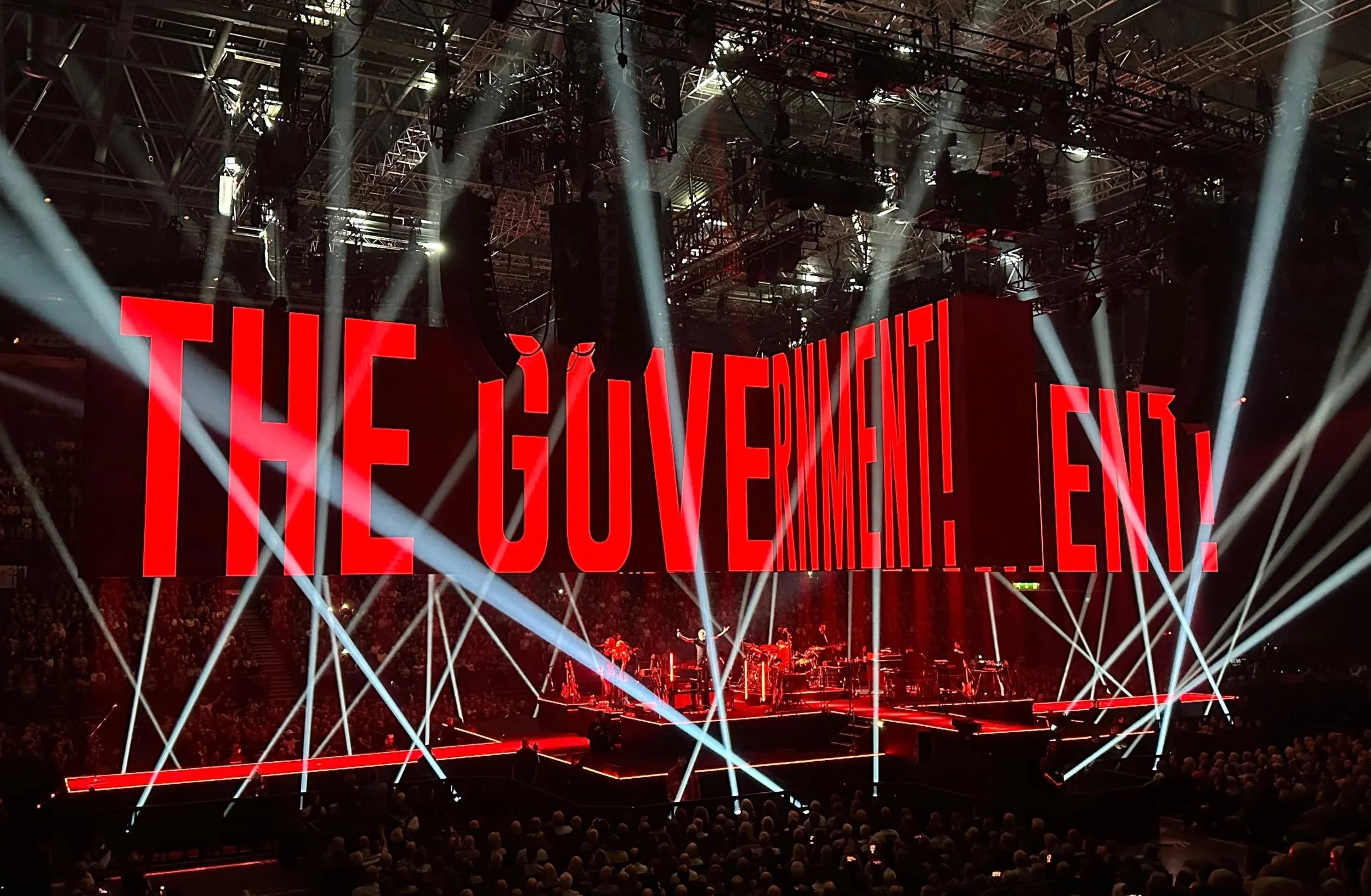 Roger Waters live on stage at Utilita Arena Birmingham with stark red LED screen projections and criss-crossing spotlight beams.