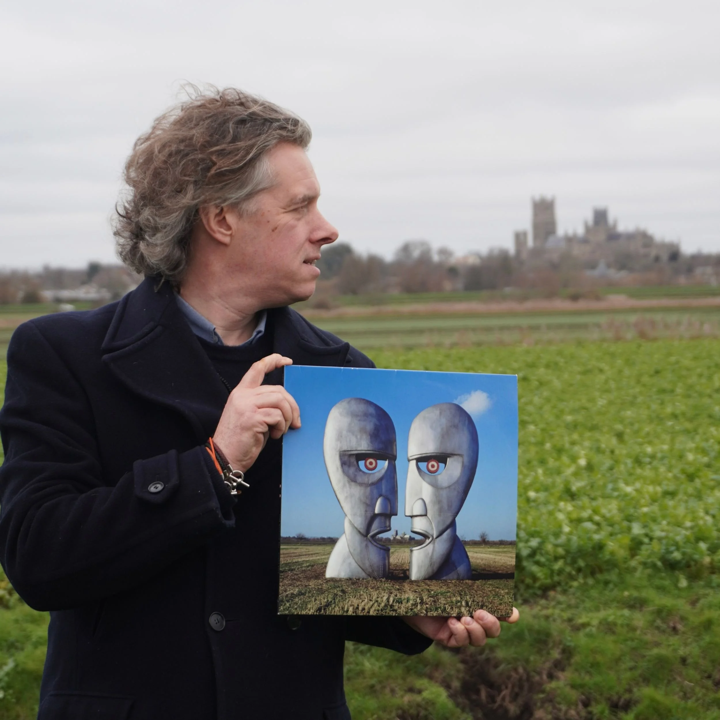 The author stands in a field in front of Ely Cathedral holding a copy of the Division Bell album cover in front of Ely Cathedral on the horizon