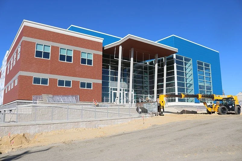 Under construction modern building with red brick and blue panels, large glass windows, and a prominent entrance with pillars, set against a clear blue sky.