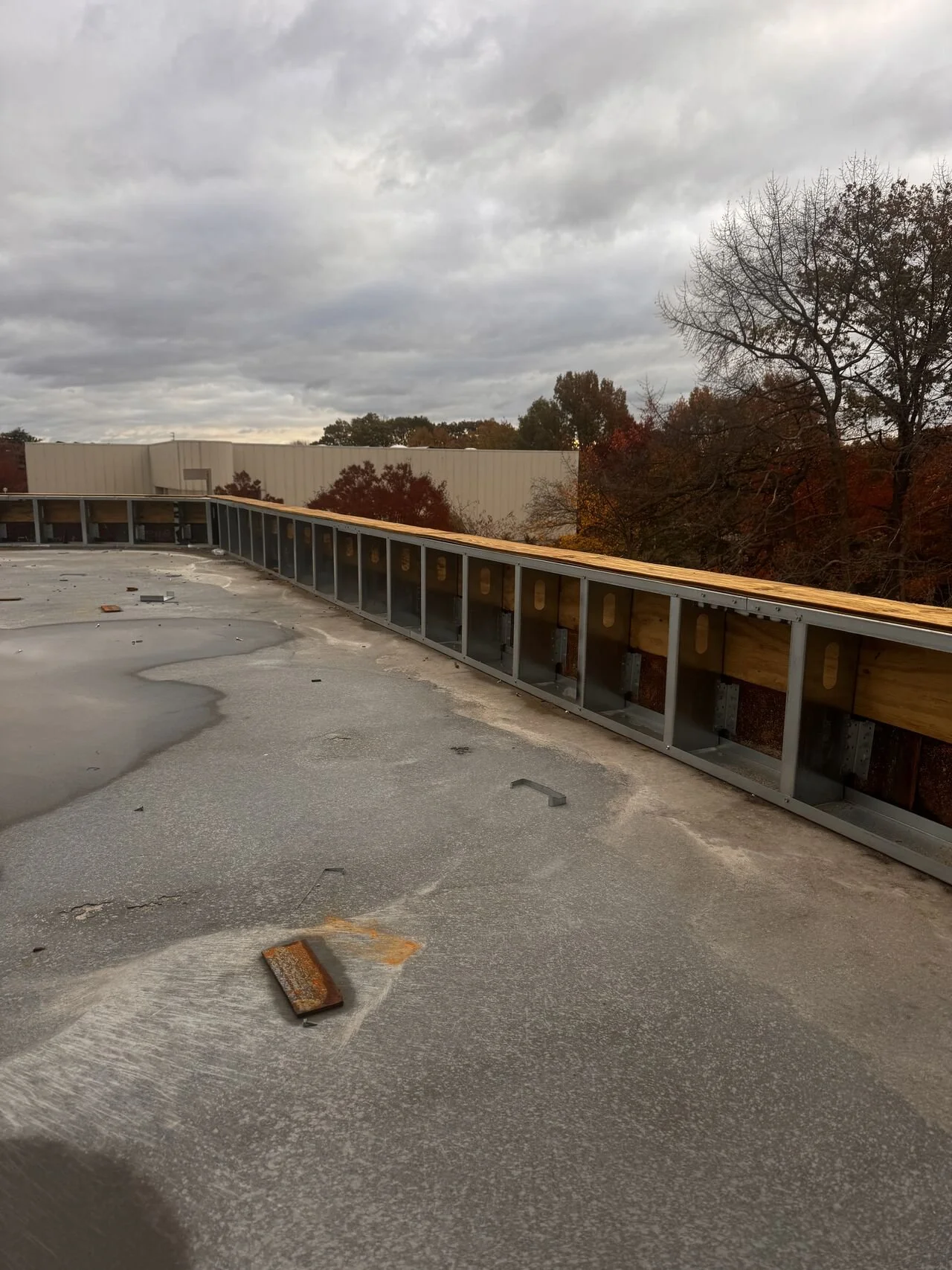 A rooftop under construction with exposed metal framing and a weathered surface, with trees and a cloudy sky in the background.