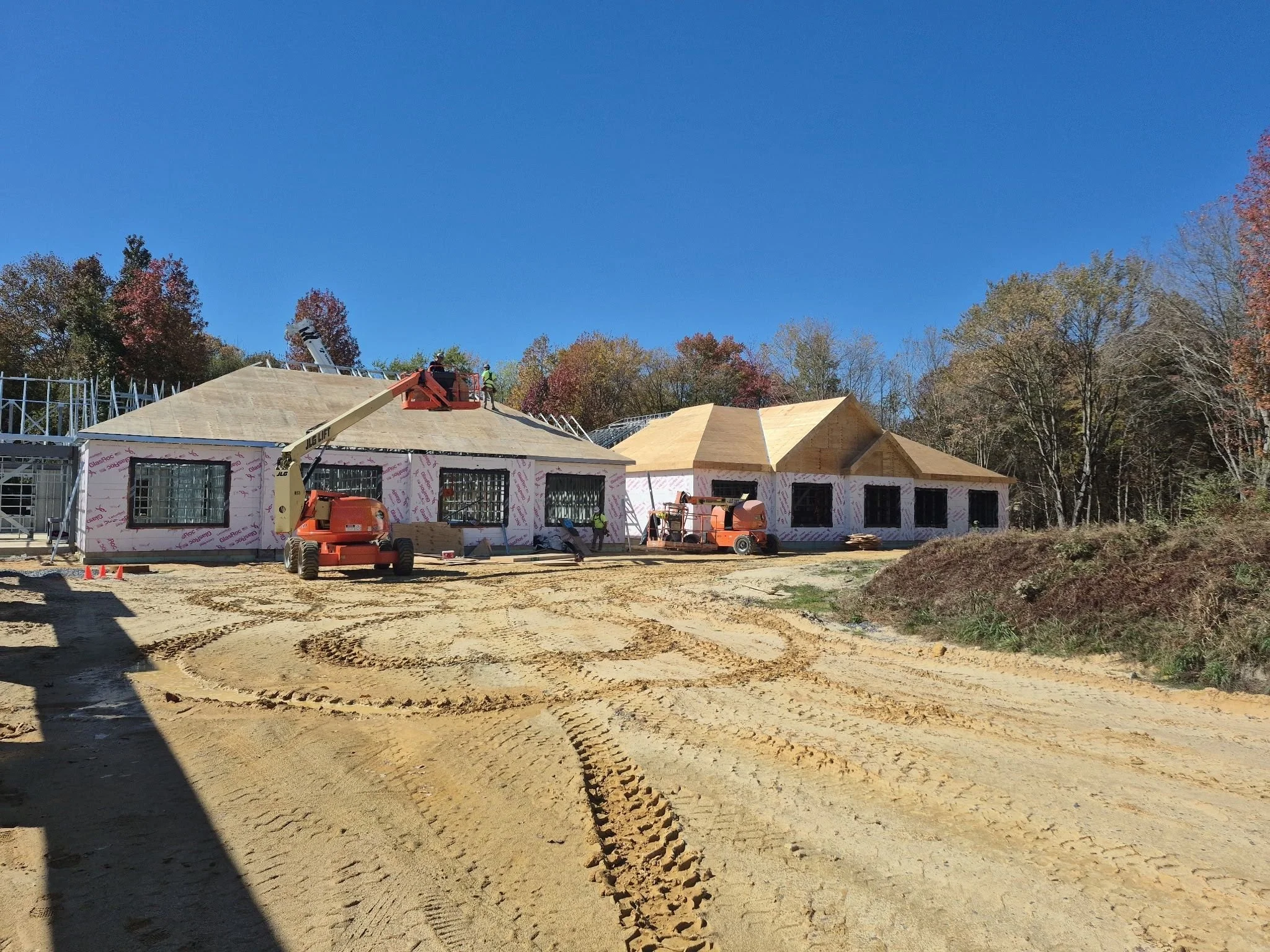 Construction site of houses with workers and machinery, dirt ground, and trees in the background under a clear blue sky.