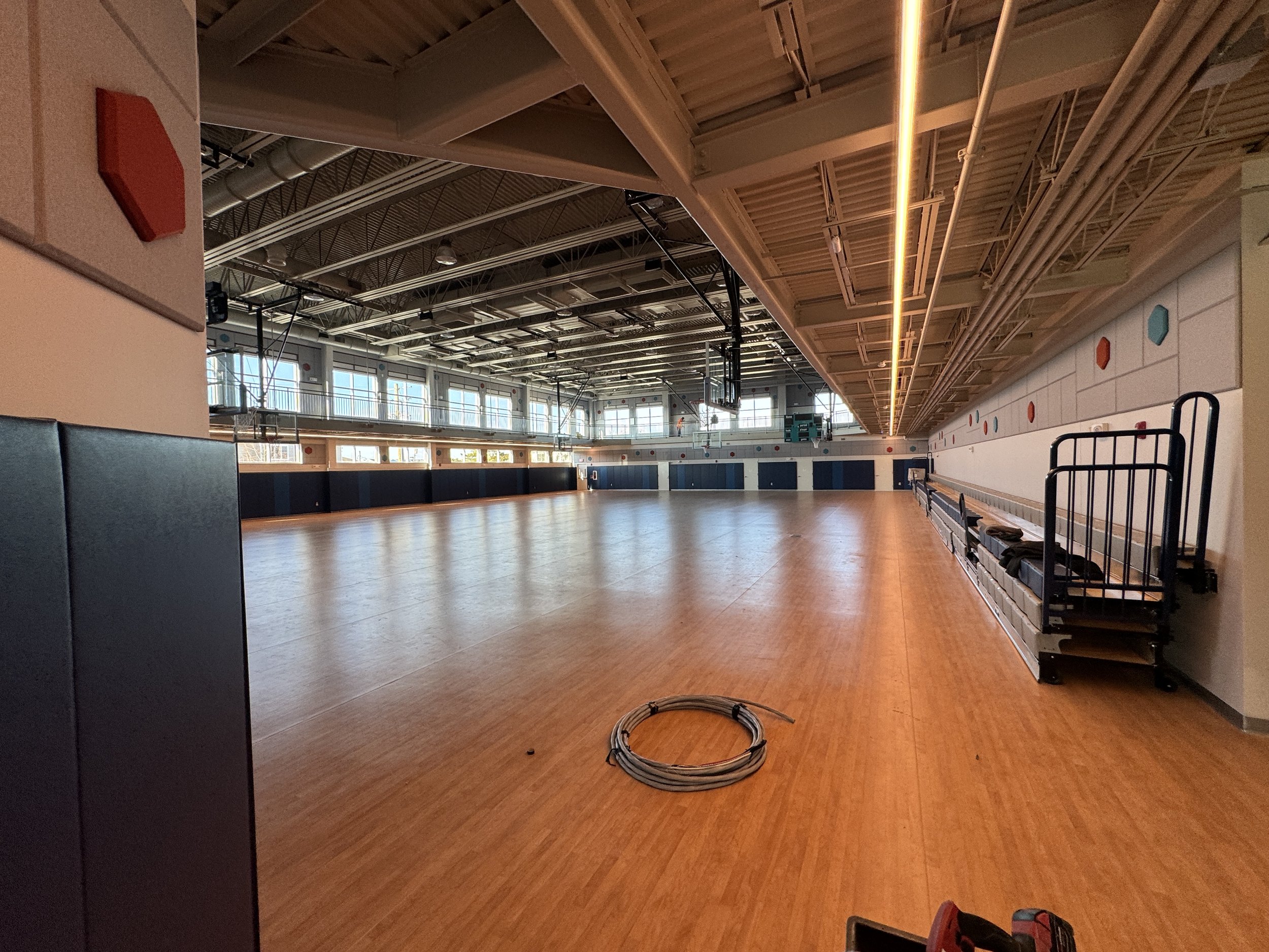 Empty indoor sports arena with a polished wooden floor, large windows, and seating along the walls. Lights and structural elements are visible on the ceiling.