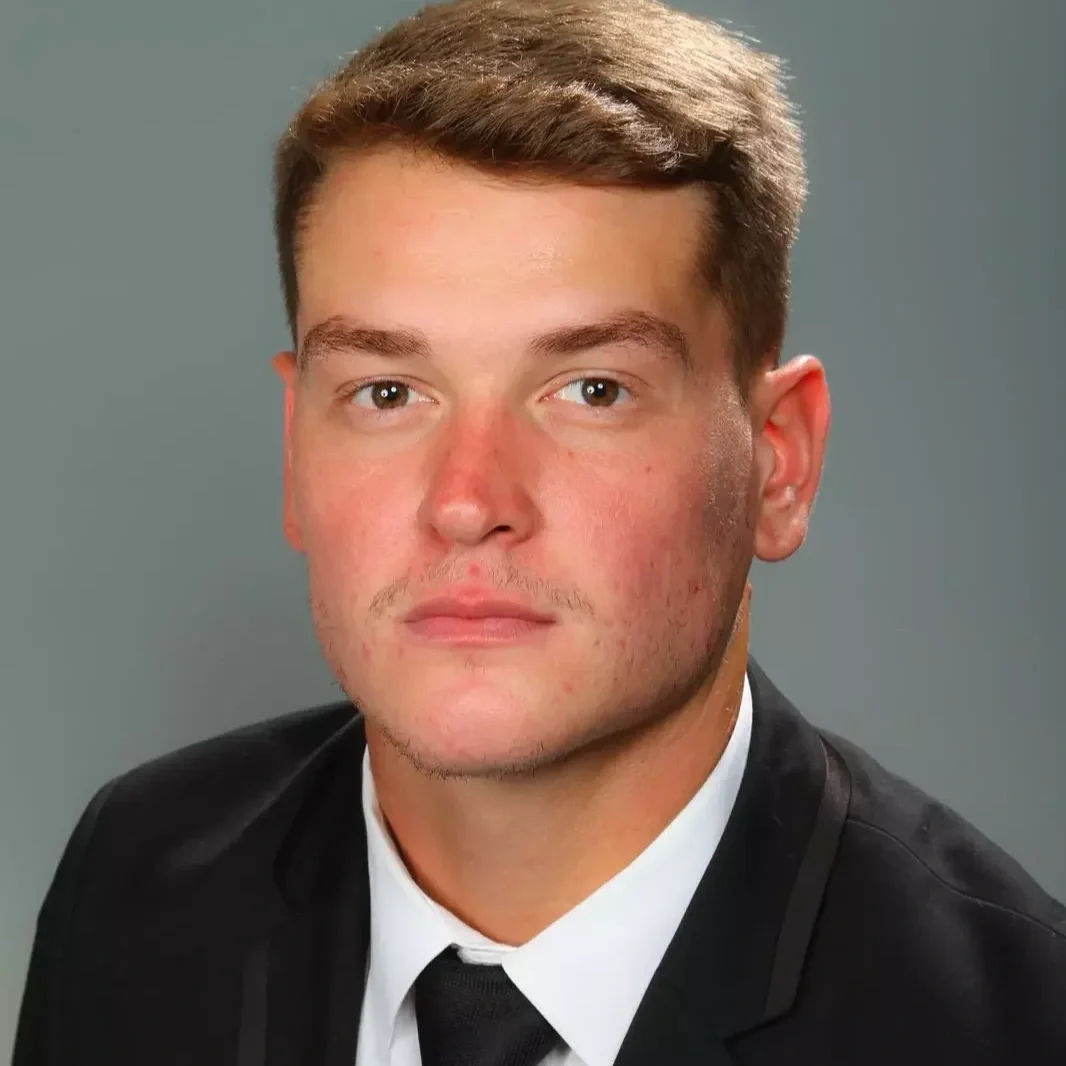 Portrait of a young man with short brown hair, fair skin, and a slight smile, wearing a black suit and white shirt against a gray background.