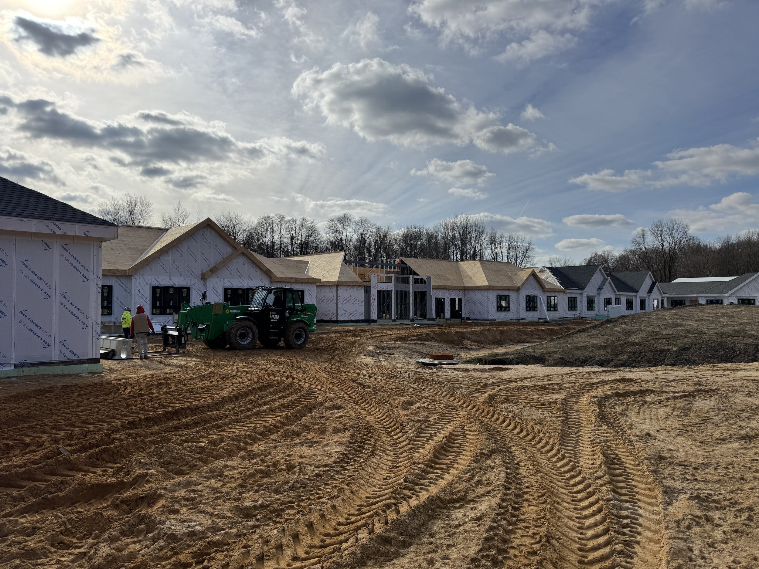 Construction site with houses under construction, construction workers, and a green skid-steer loader on dirt ground, with a partly cloudy sky.