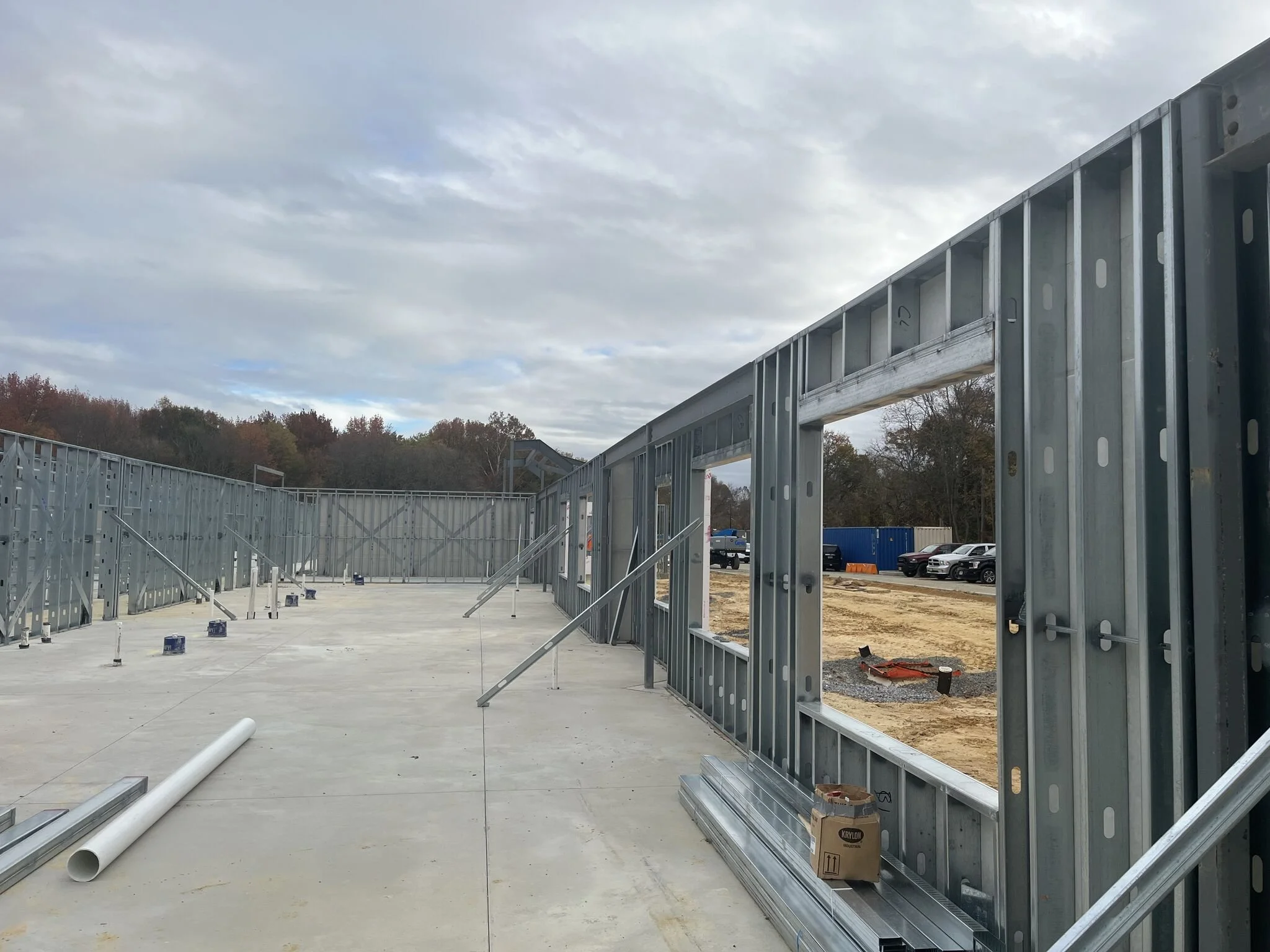 Construction site with metal framing and a concrete floor, with trees and parked cars in the background under a cloudy sky.