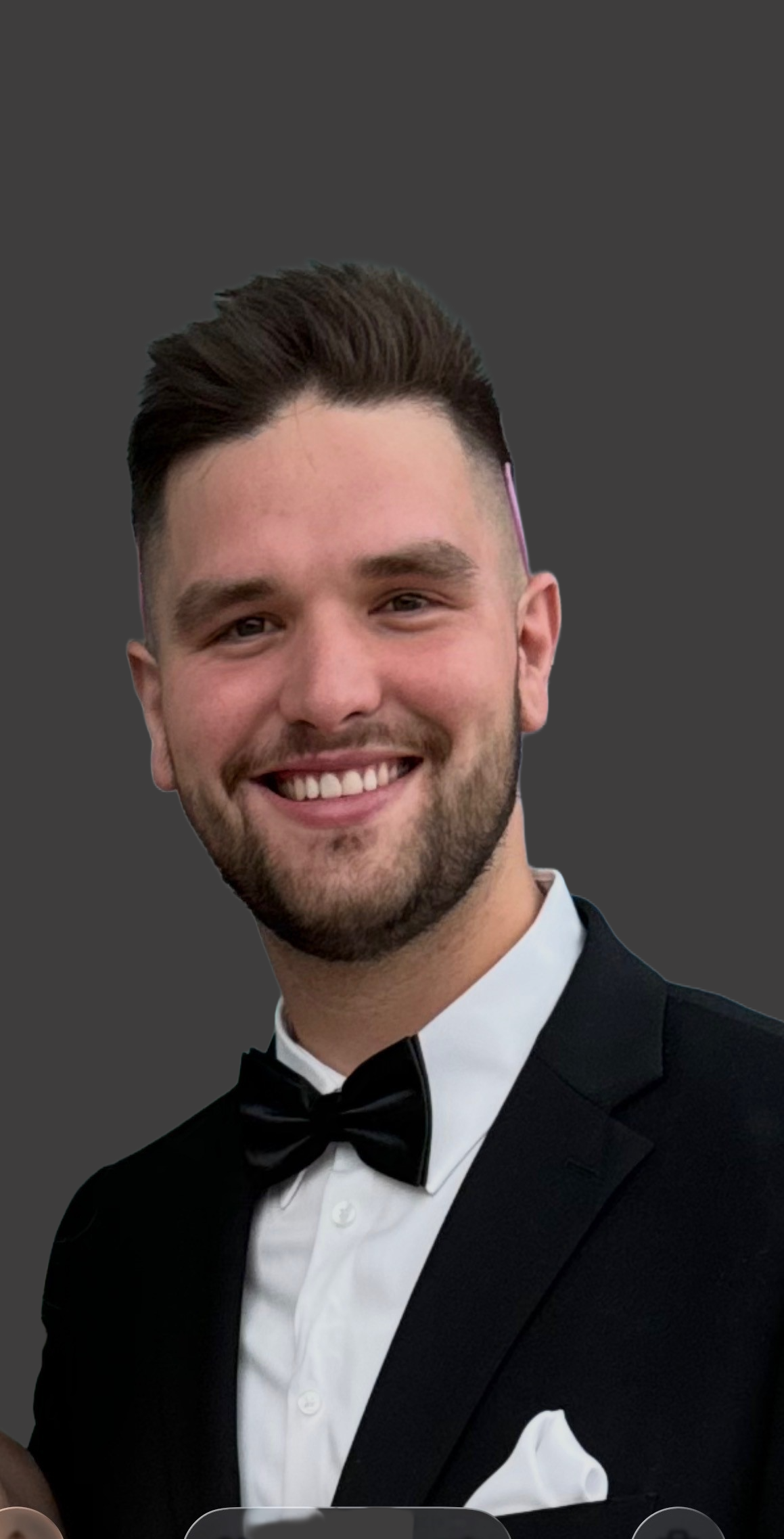 A smiling young man with a beard and styled hair wearing a tuxedo with a black bow tie and white shirt, against a dark background.
