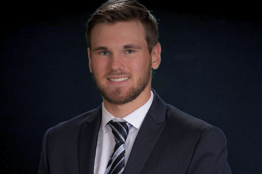 Portrait of a young man with short brown hair and a beard, wearing a dark suit with a white shirt and a patterned tie, smiling in front of a dark background.