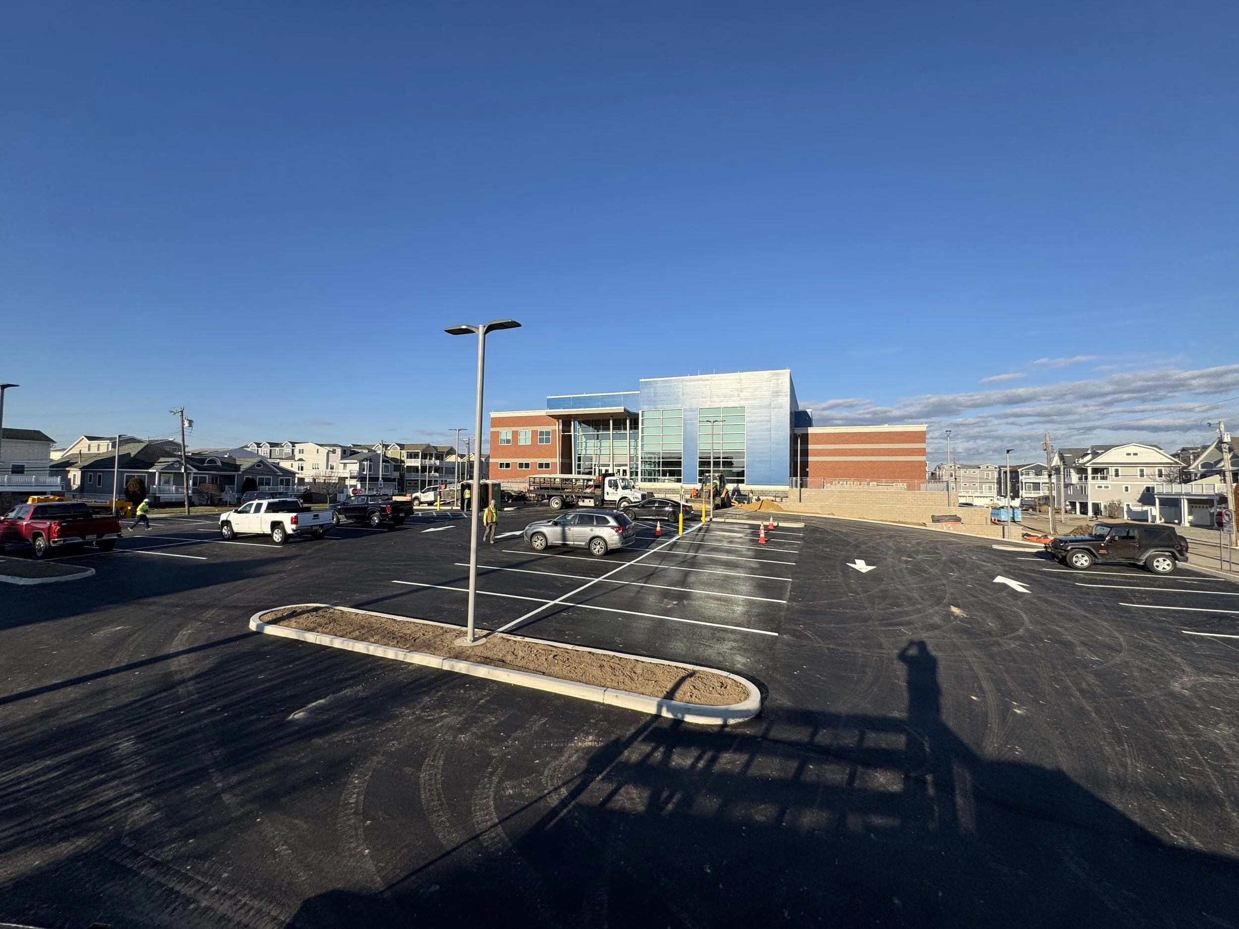 Empty parking lot next to a modern building with construction in progress, and residential houses in the background under a clear blue sky.