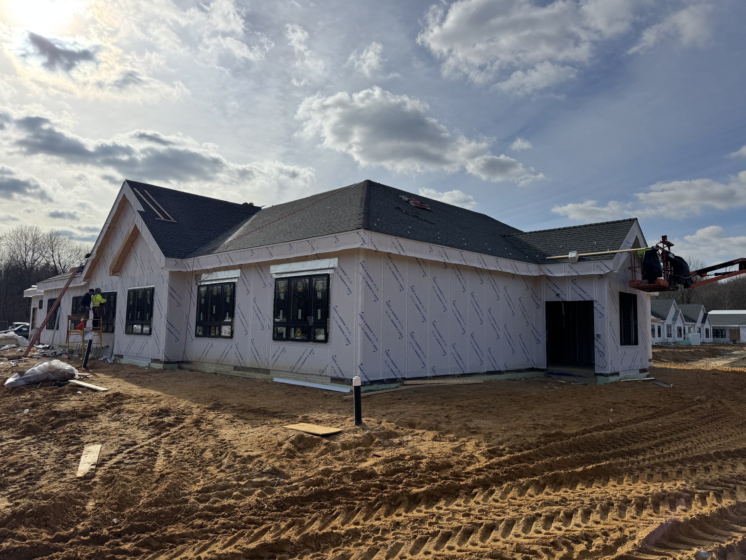 A house under construction with workers installing the roof, surrounded by dirt and construction equipment, under a partly cloudy sky.