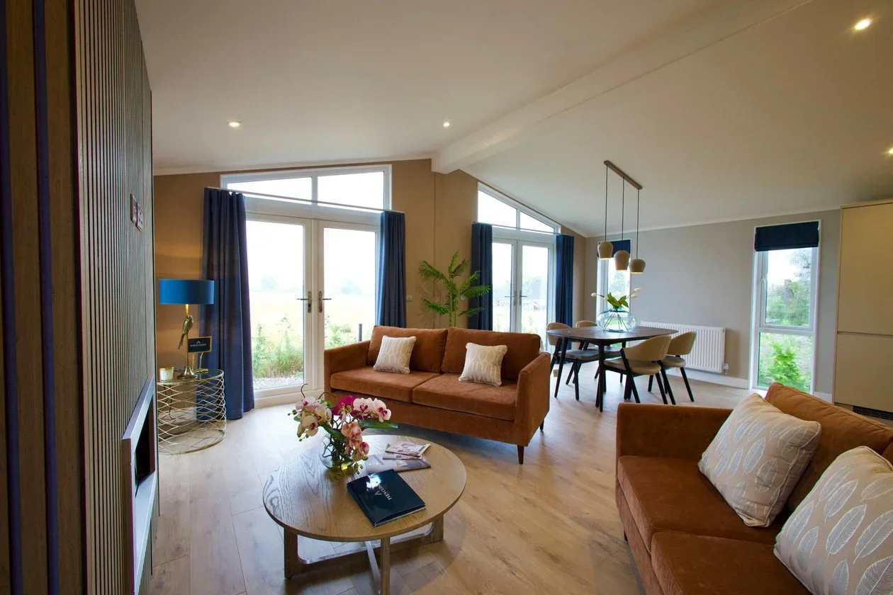 Living room with two brown sofas, a round coffee table with flowers and a book, large windows with blue curtains, a dining table with chairs, a blue floor lamp, and a potted plant.