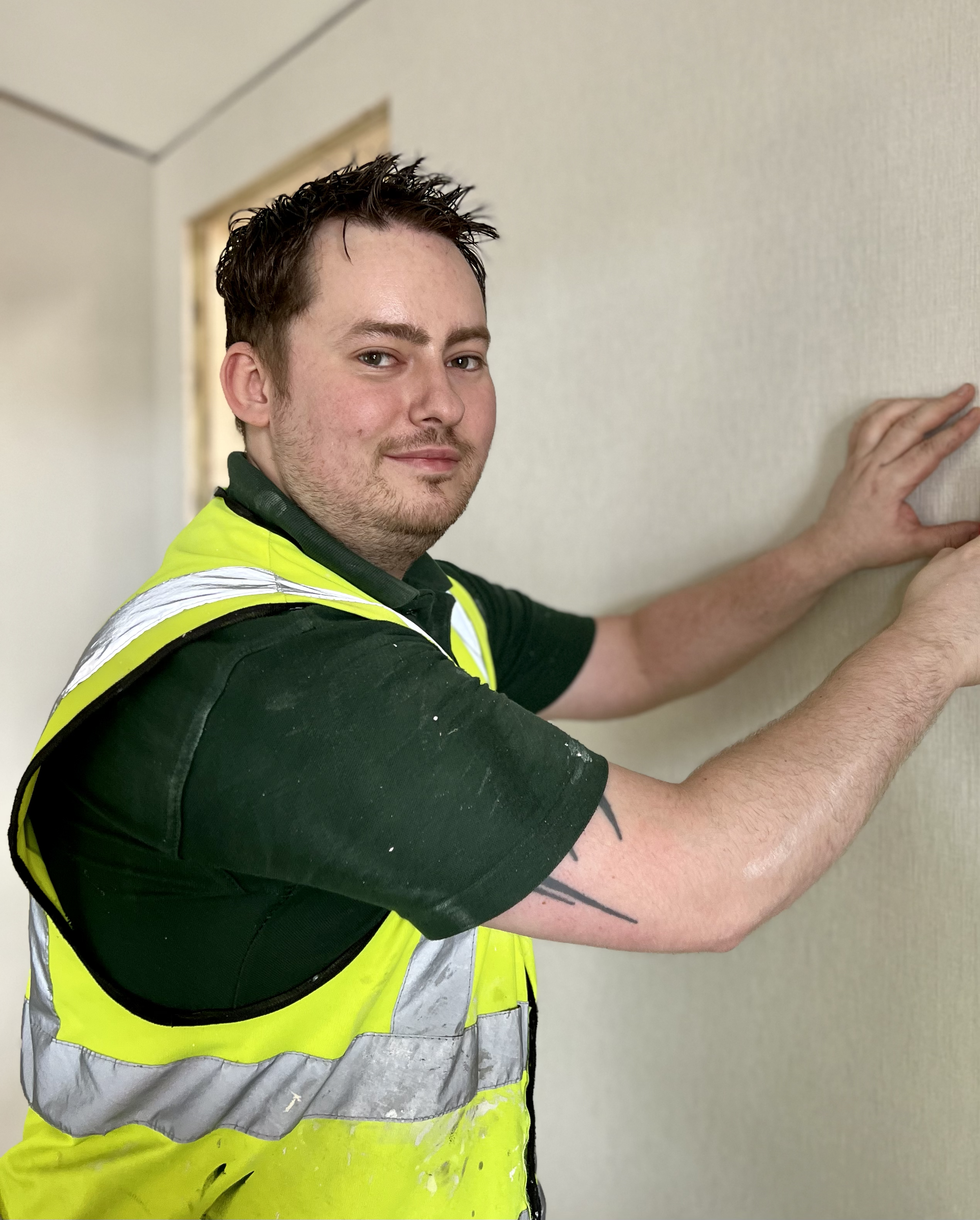 A man in a black shirt and a yellow safety vest is pressing his hands against a beige wall, standing in a hallway.