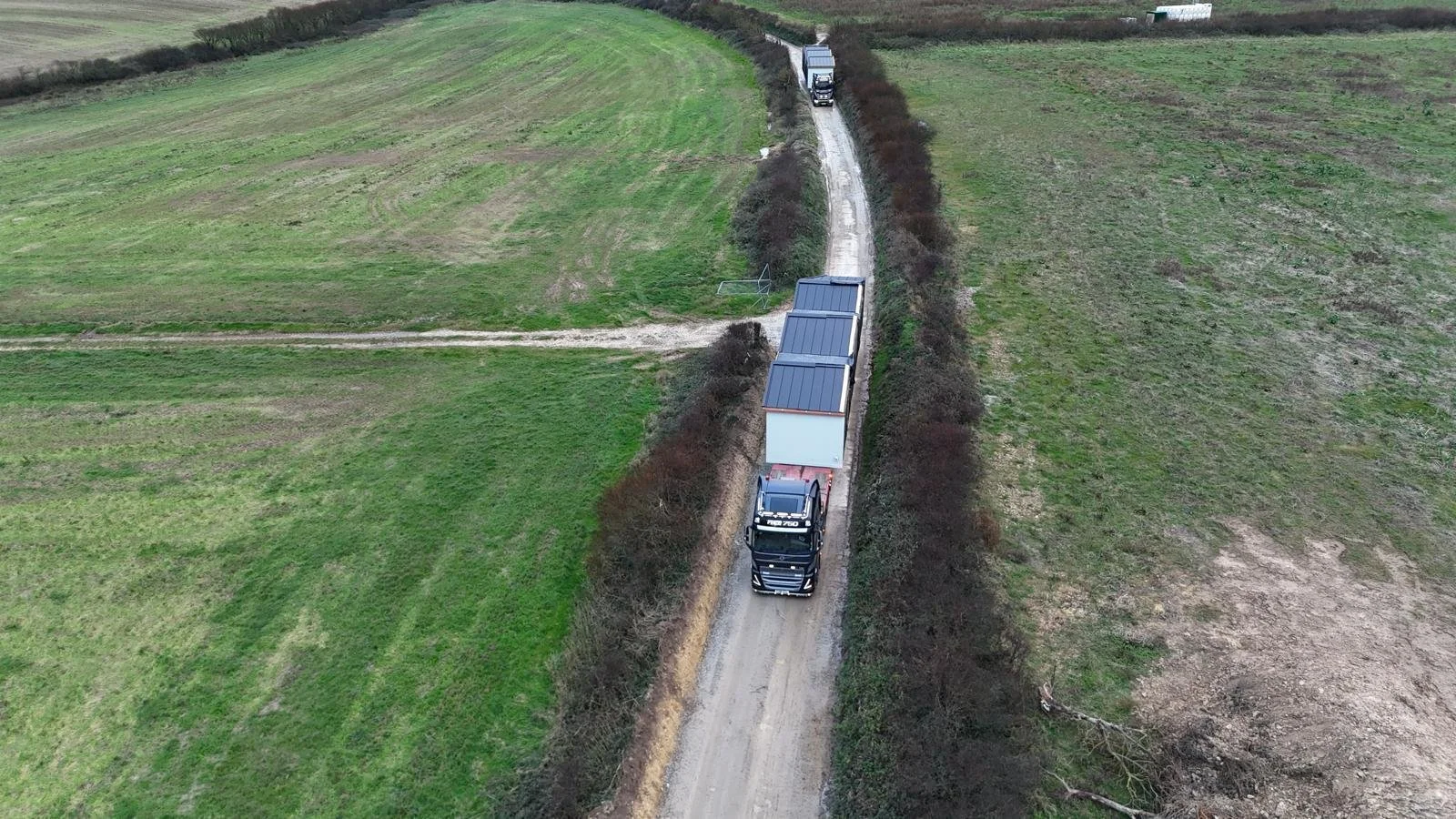 Aerial view of a large truck with solar panels on its roof traveling on a narrow dirt road through green fields, bordered by bushes.