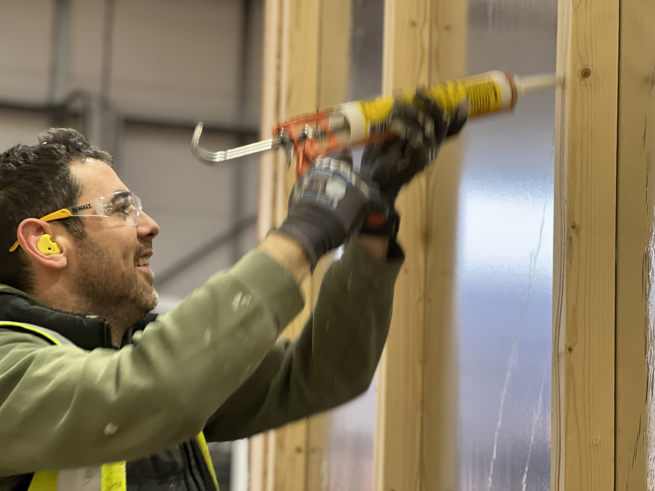 A man wearing safety glasses, ear protection, gloves, and a green jacket is using a caulking gun to apply caulk to a wooden wall in an indoor construction setting.