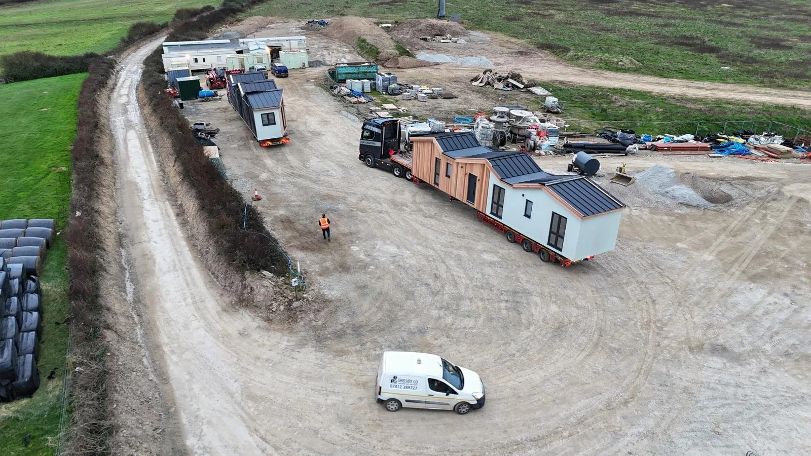 Construction site with a moving trailer carrying a modular house, construction workers, and building materials scattered around.
