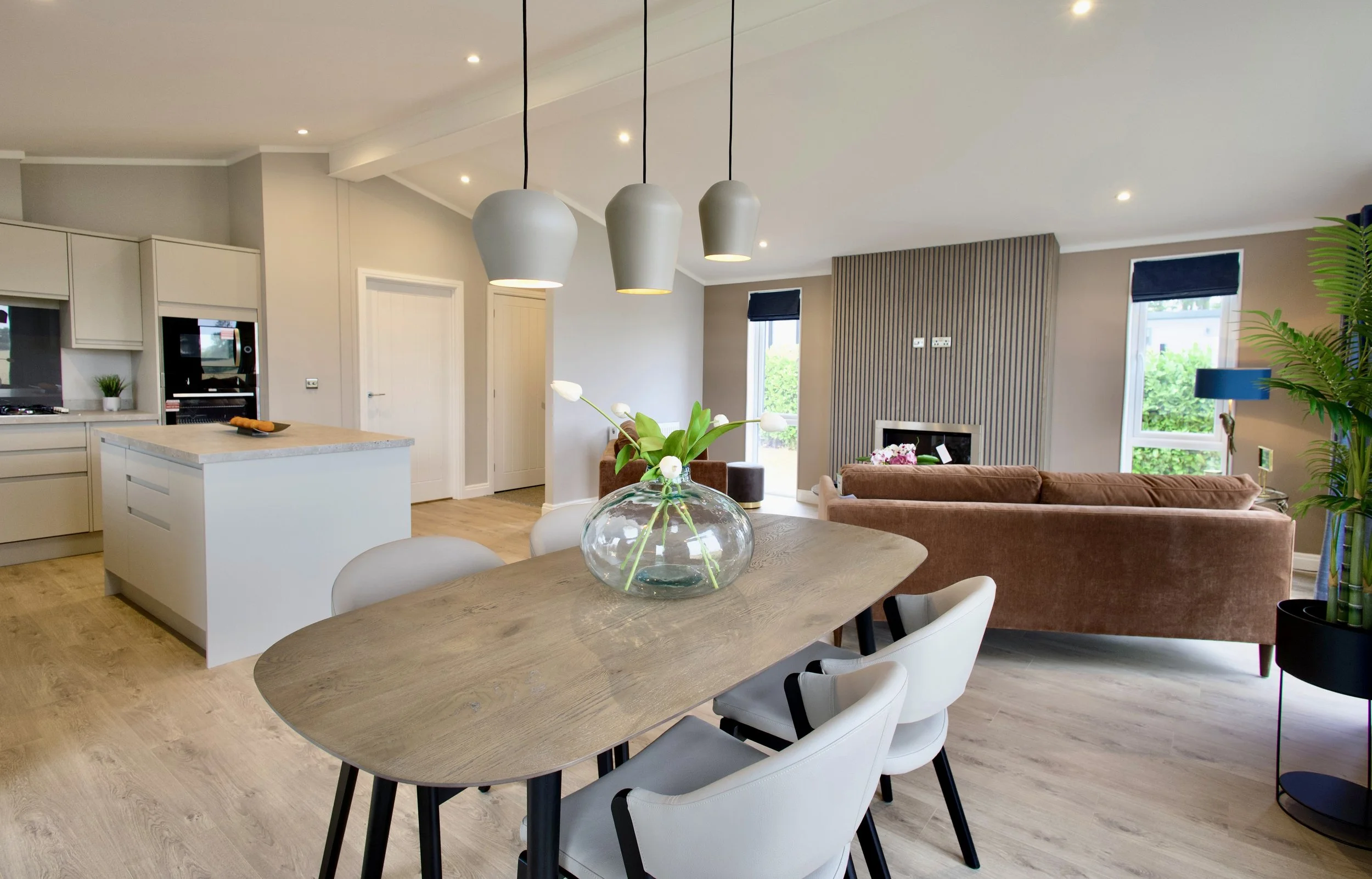 Open-concept living and dining room with modern decor, a wooden dining table with white chairs, a large glass vase with flowers, a sofa, and a fireplace, illuminated by natural light through windows and pendant lights