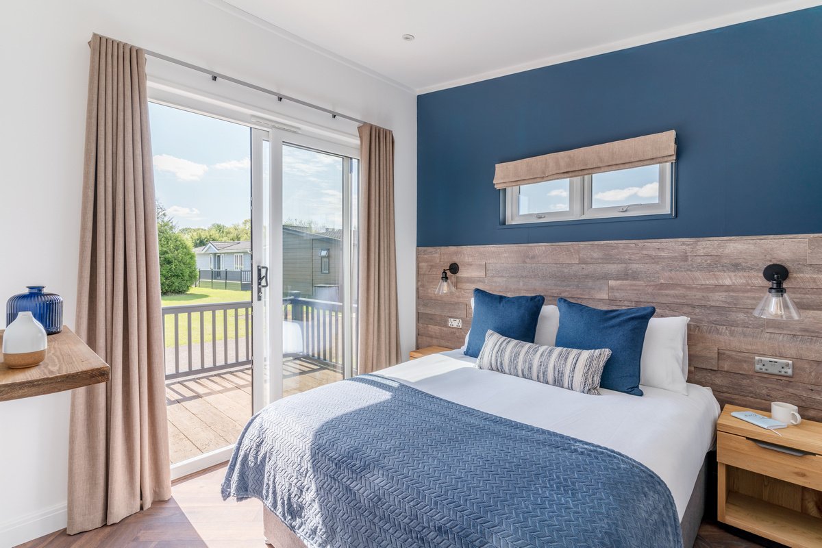 A bedroom with a sliding glass door open to a small balcony, wooden flooring, a bed with blue pillows and a quilt, beige curtains, and a wooden headboard with small wall-mounted lamps.