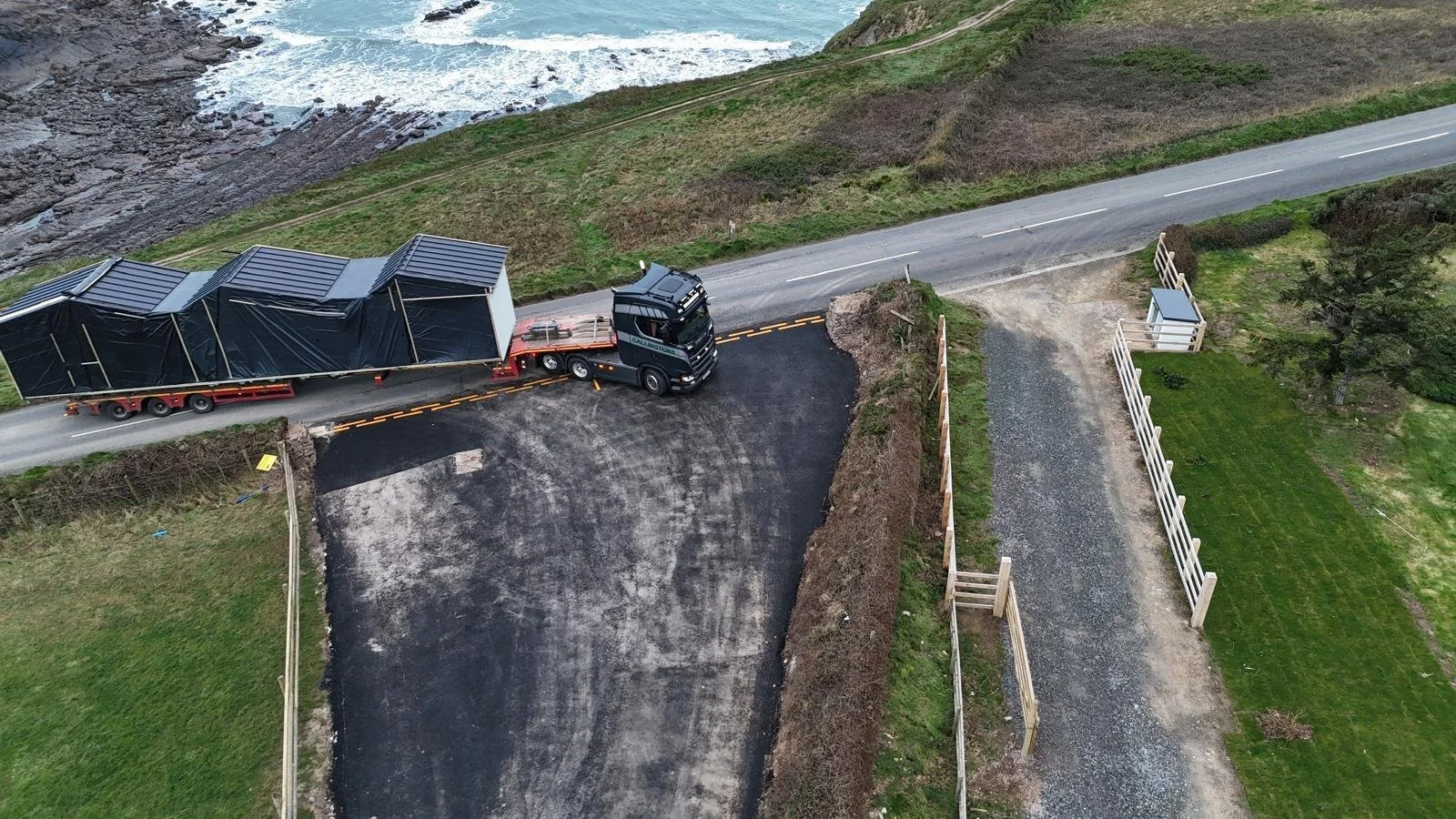 A large truck with a trailer carrying black cargo is parked on a dirt and asphalt driveway by a winding coastal road. The scene shows a hillside with grass and bushes, a gravel path bordered by wooden fencing, and the ocean in the background.