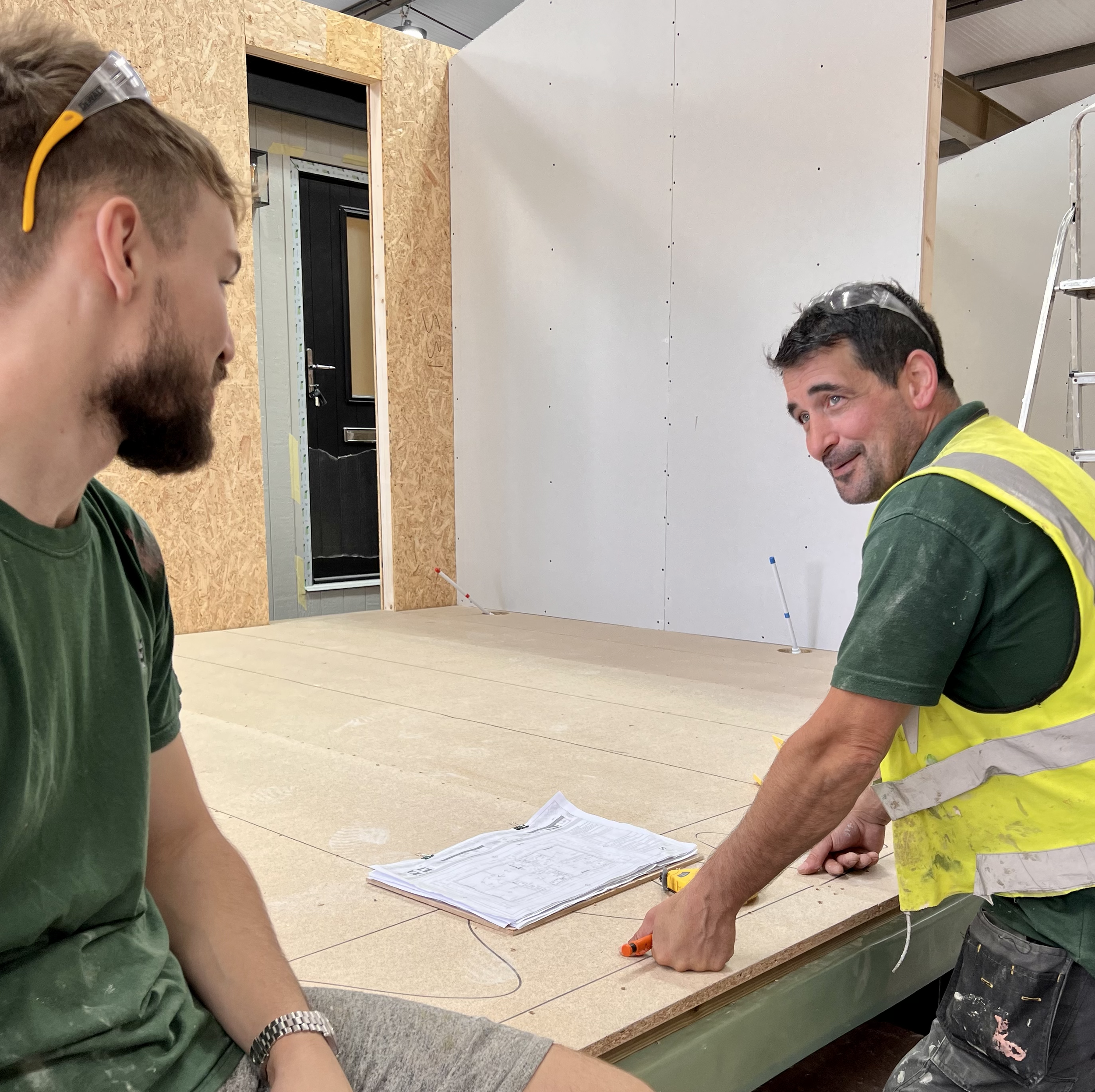 Two construction workers talking on a construction site, one wearing a yellow safety vest and the other wearing a green shirt, with construction plans on the table.