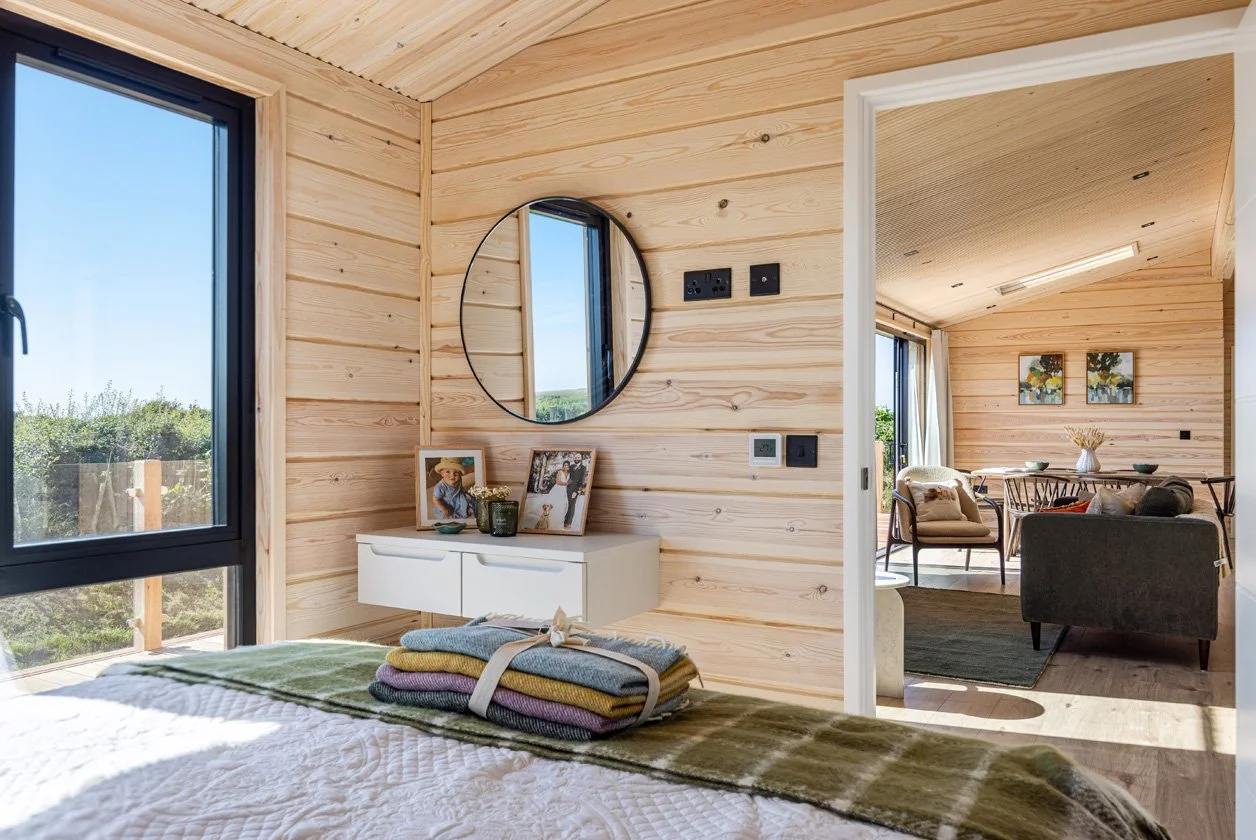 Bedroom with wooden walls, a bed with folded towels, a mirror, and a white dresser with picture frames and decor, with a view of the outdoors through a large window. An adjoining living room with seating and framed artwork is visible.
