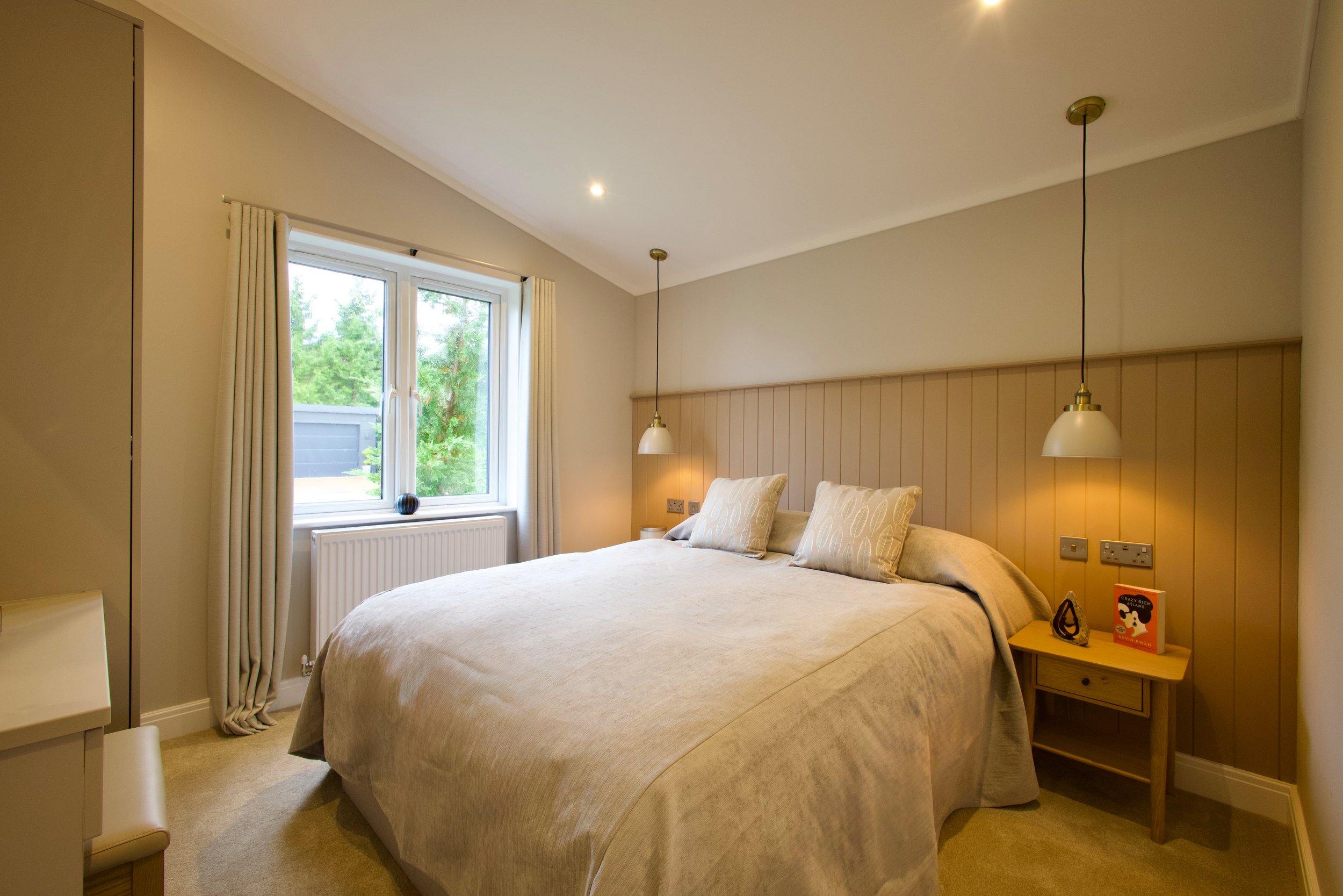 A cozy bedroom with a large bed, beige bedding, and three matching pillows. Two pendant lights hang above the nightstand, which holds a colorful book, a small sculpture, and a decorative item. There is a window with cream-colored curtains revealing a