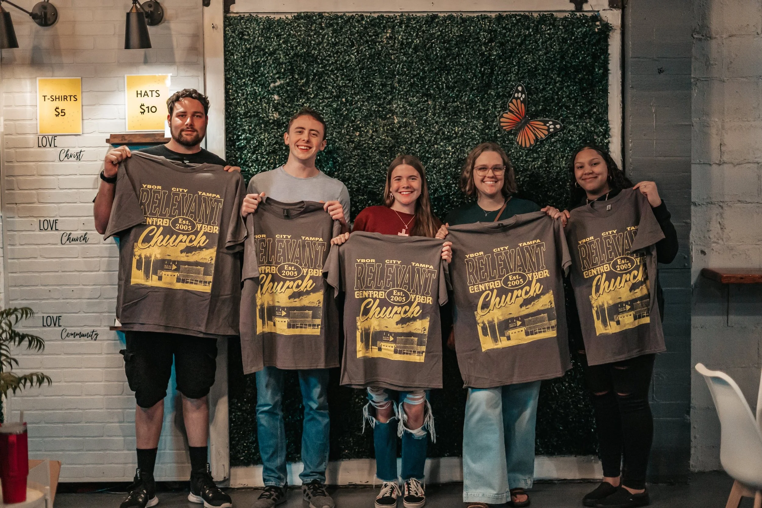 A group of five people holding gray T-shirts with a graphic design that says "Relvant Church" and lists "Ybor City," "Tampa," and "Est. 2005." They are standing in a room with a decorative green leafy wall, queen butterfly wall art, and signs on the wall indicating merchandise prices for T-shirts and hats.