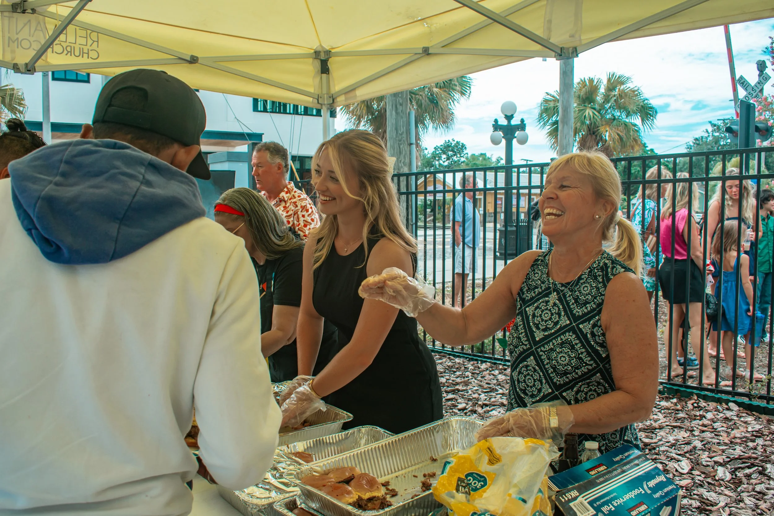 Smiling woman serving food at outdoor event under yellow canopy, with people and palm trees in the background.