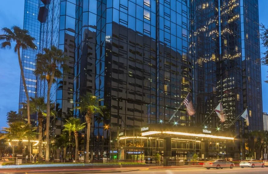 Night view of a tall glass skyscraper with the Hilton hotel entrance at street level, decorated with string lights and flags, with palm trees lining the sidewalk.