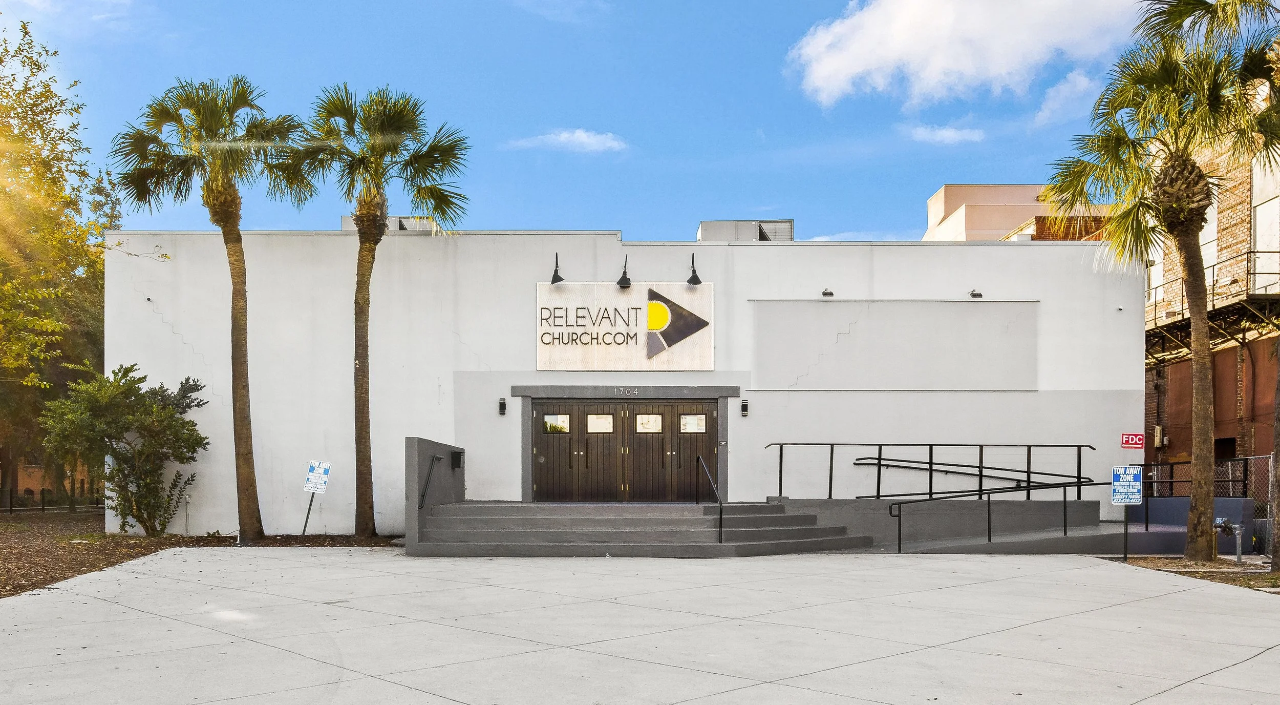 Front view of a white church building with palm trees on either side, a dark door, and signage reading 'RELEVANT CHURCH.COM' under a bright blue sky with some clouds.