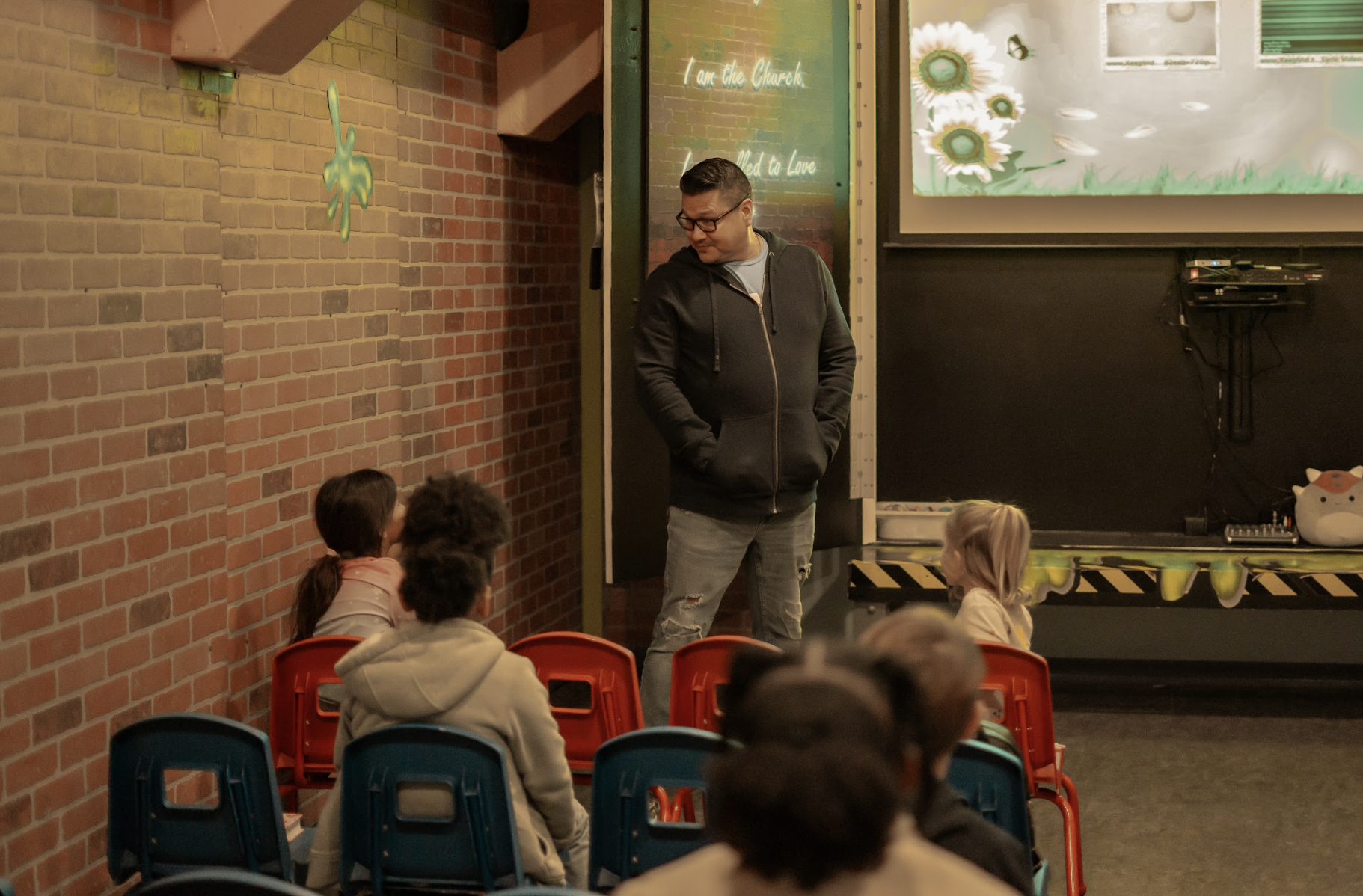 A man standing in front of a group of children seated on colorful chairs listening to him indoors, with a brick wall and a screen in the background.