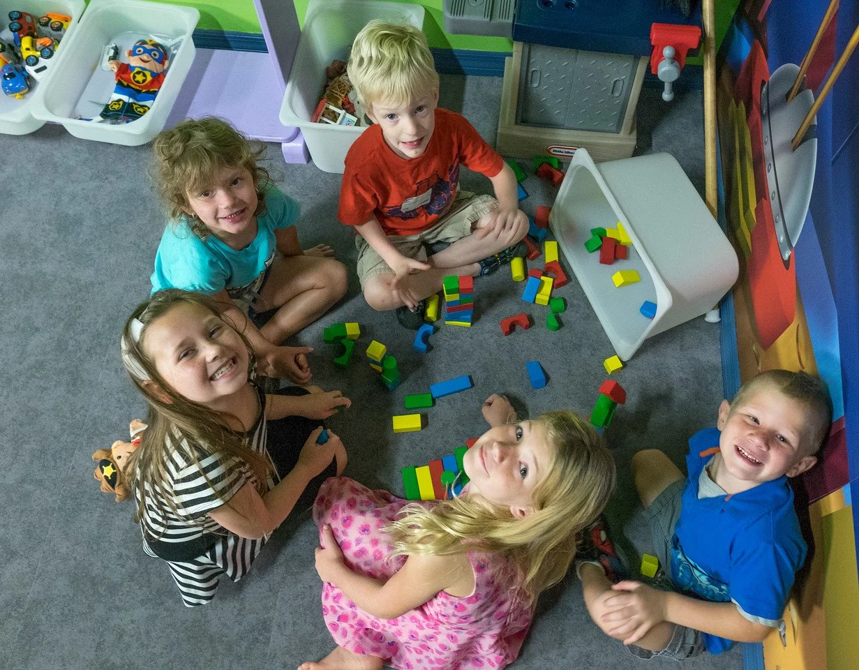 Group of five children playing with colorful building blocks on a gray carpet in a playroom.