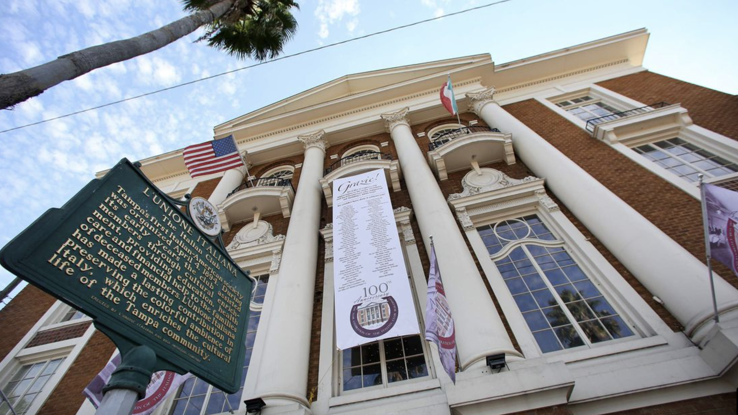 Historical building with white columns and brick facade, displaying flags of the United States and Italy, and banners celebrating its 100th anniversary.