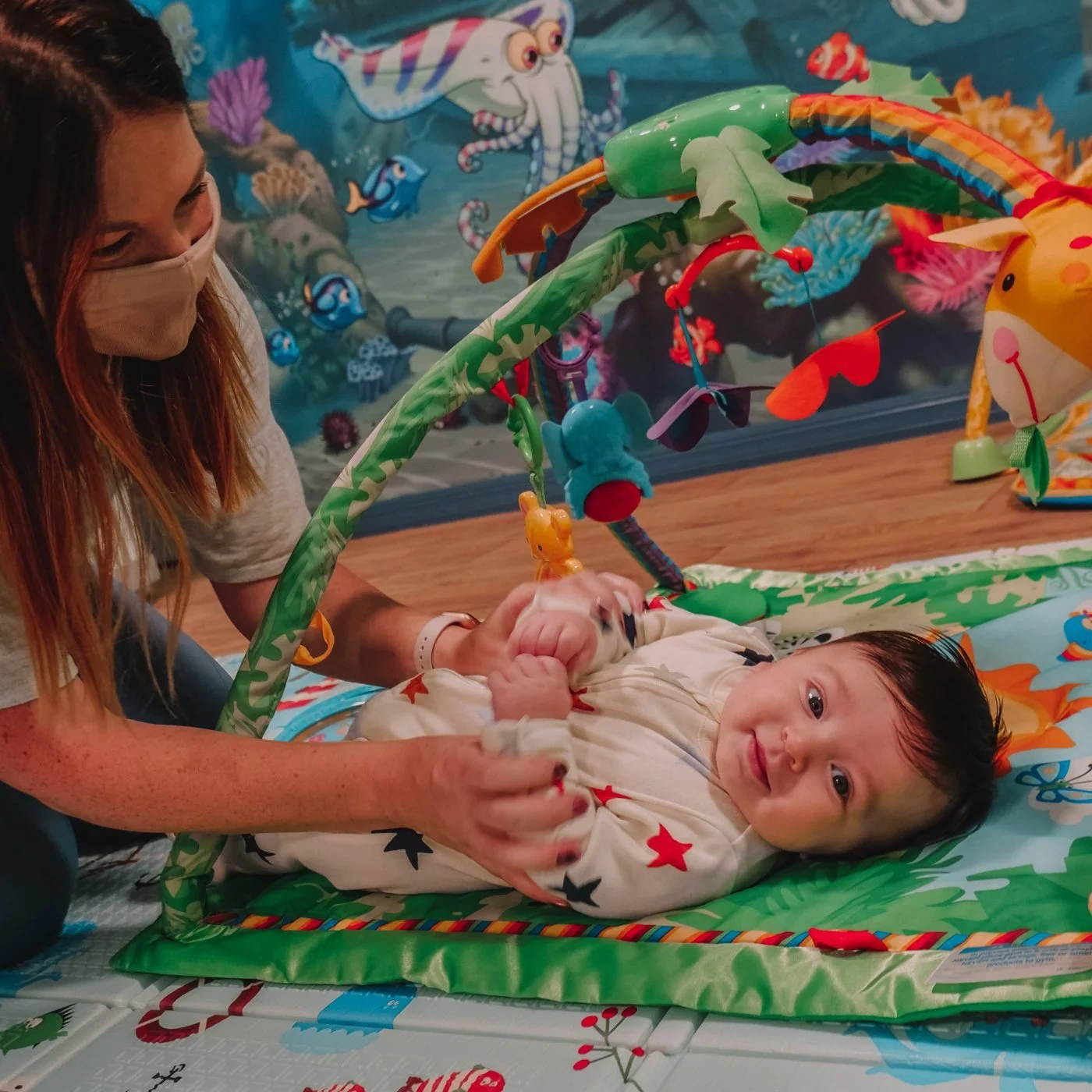 A woman wearing a mask is holding a baby lying on a colorful play mat with an overhead toy arch. The background features an ocean-themed wall mural with fish and sea creatures.
