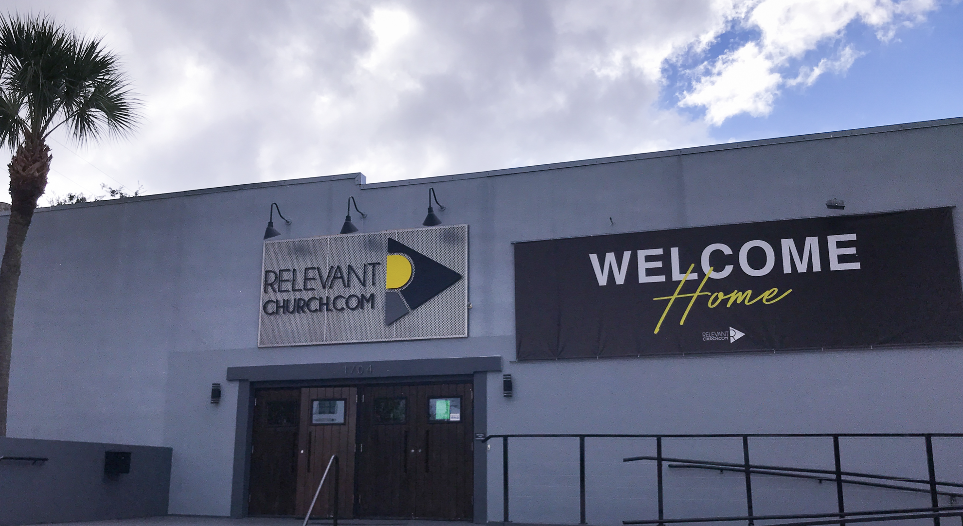 The exterior of a building with two signs; one reading 'ReRelevant Church @CM' with a logo and the other reading 'Welcome Home' with the church's name and logo.