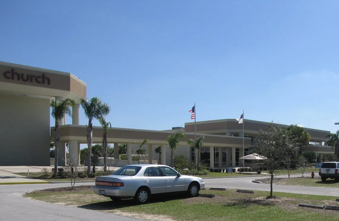 A church building with palm trees, a parking lot with a car, and flags in the background, under a clear blue sky.