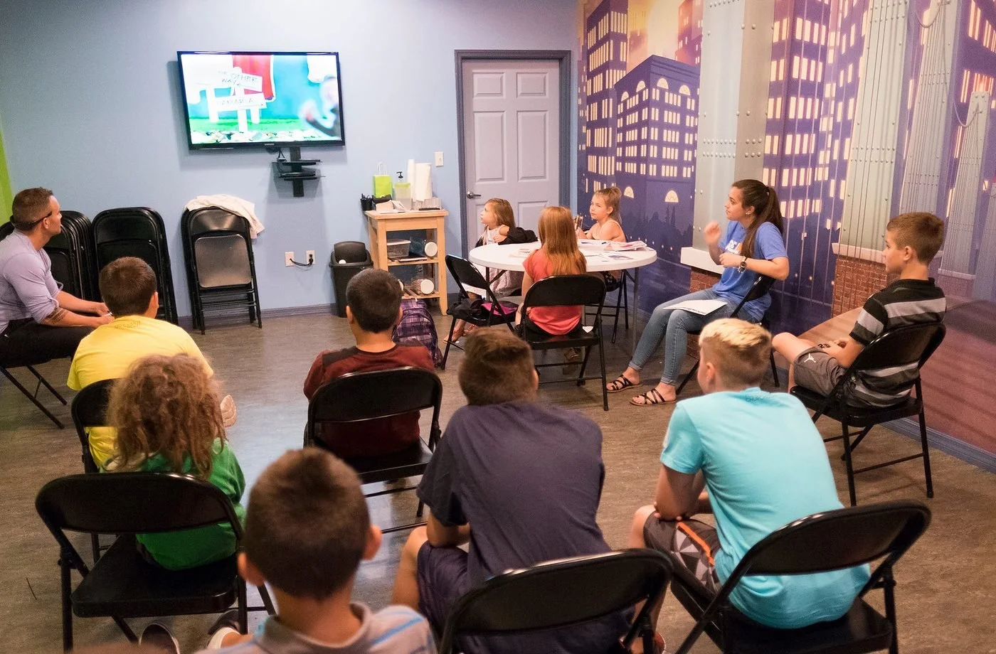 A group of children and a woman are watching a presentation on a TV screen in a classroom or meeting room. The room has a cityscape mural on one wall and is equipped with chairs, a small table, and a cart with supplies.
