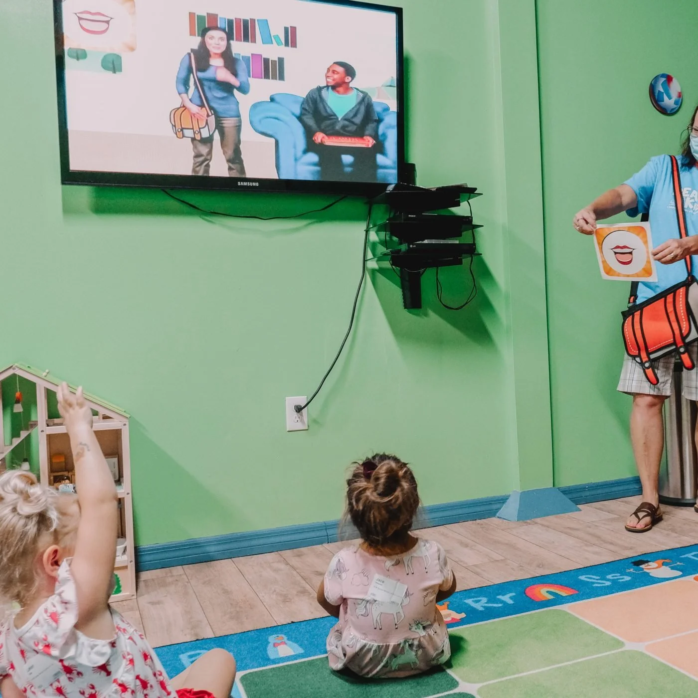 Children sitting on the floor in front of a television screen, one child raising their hand, in a brightly colored room, engaging in an activity with a woman holding a sign with a smiling mouth illustration.