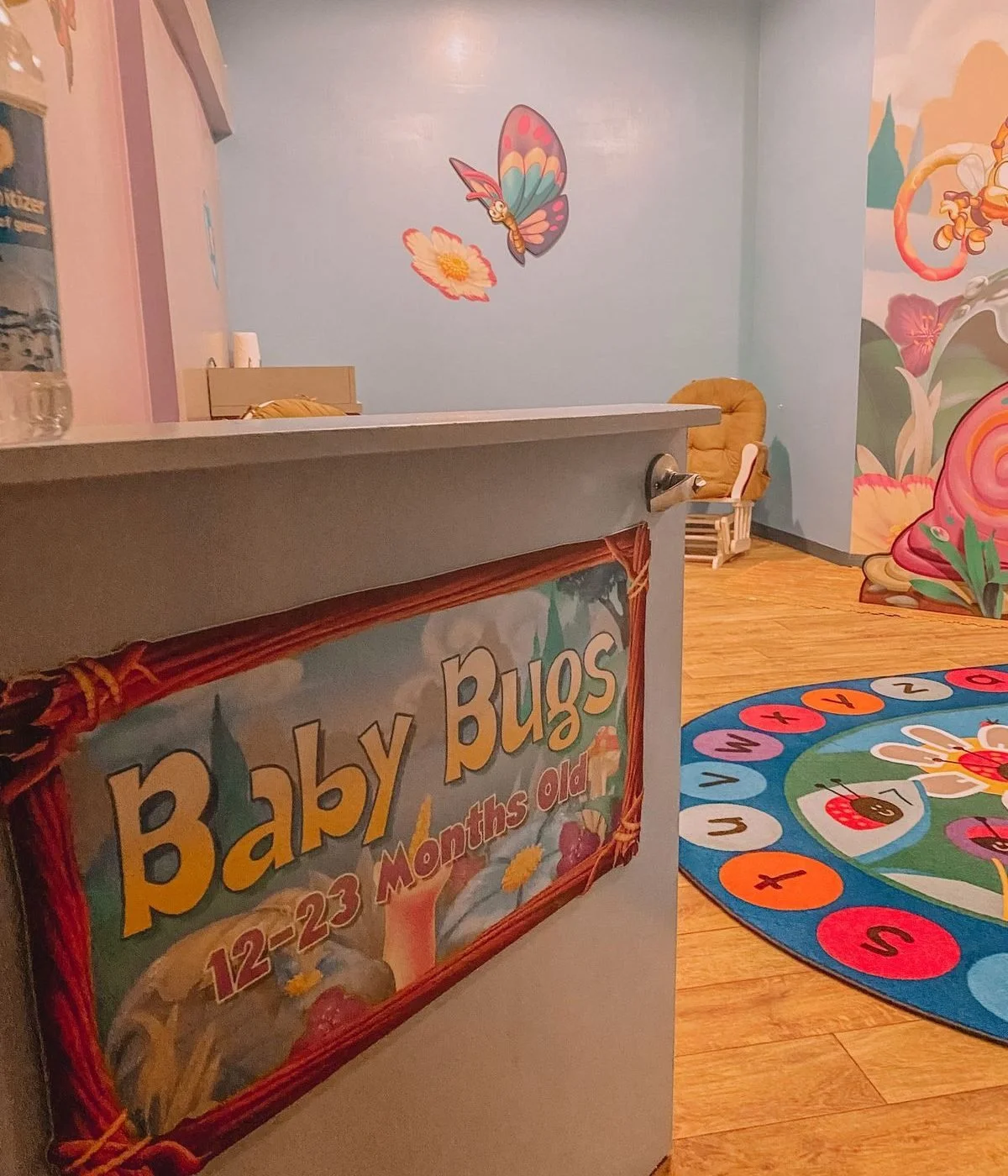 A colorful indoor children's play area with a sign that reads 'Baby Bugs 12-23 Months Old'. The area has a painted wall with a butterfly and flower mural, a multicolored hopscotch rug, and a cozy chair in the background.