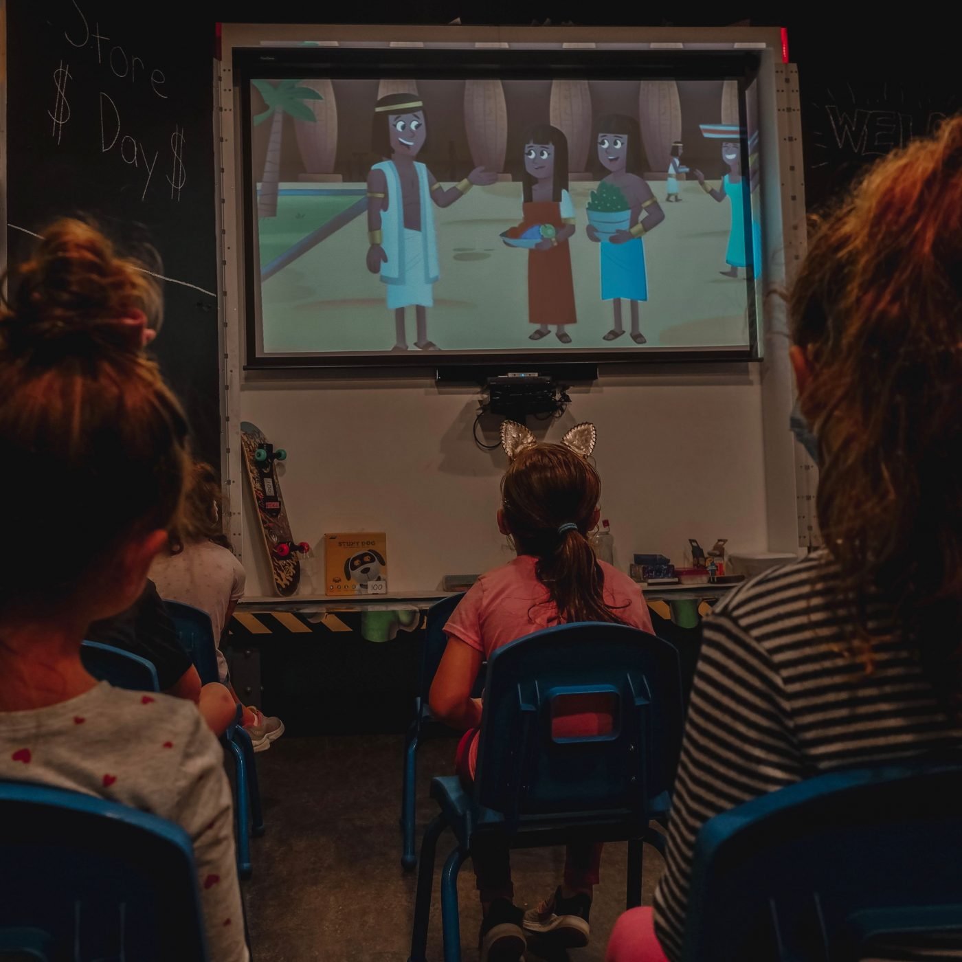 Children watching a cartoon about a beach or tropical setting on a screen in a classroom.