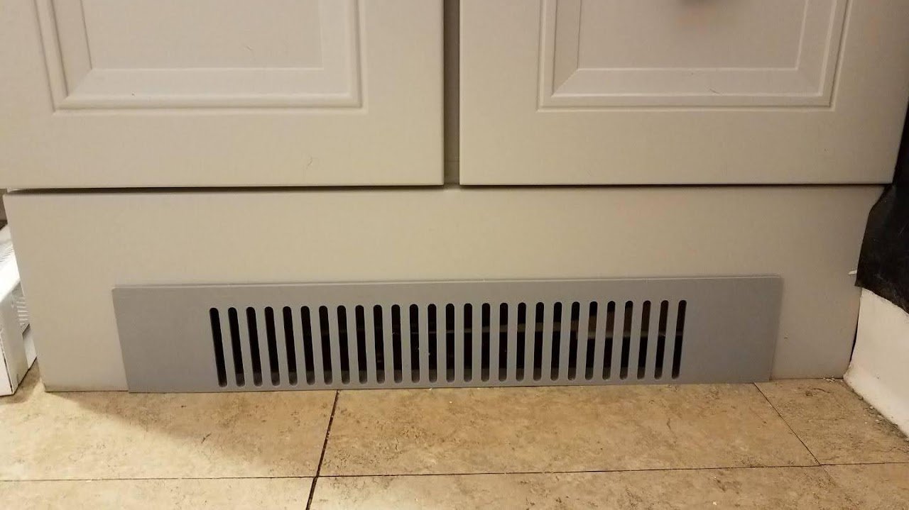 A beige colored kitchen cabinet with a gray vent below it. The vent has vertical slats and is placed on a tiled floor.