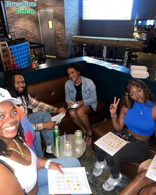 Four young women and one young man sitting in a booth at a bar or restaurant, smiling and posing for the camera. The women are holding menus and drinks, with one making a peace sign gesture. The background features brick walls, bar tables, and a large screen or window.