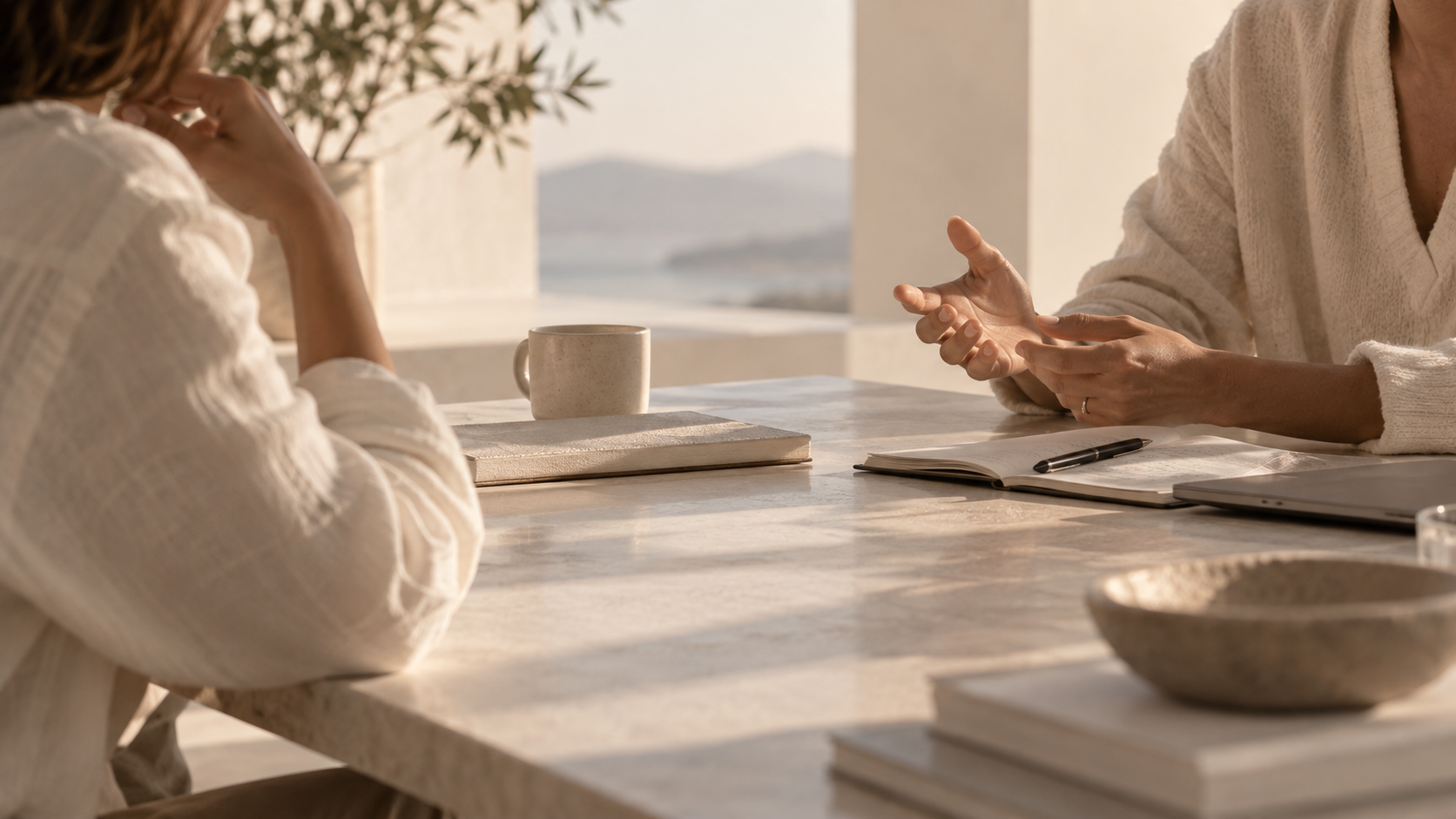 Two women having a discussion at a marble table with notebooks, a pen, and a coffee mug, in a bright room with a scenic view of mountains in the background.