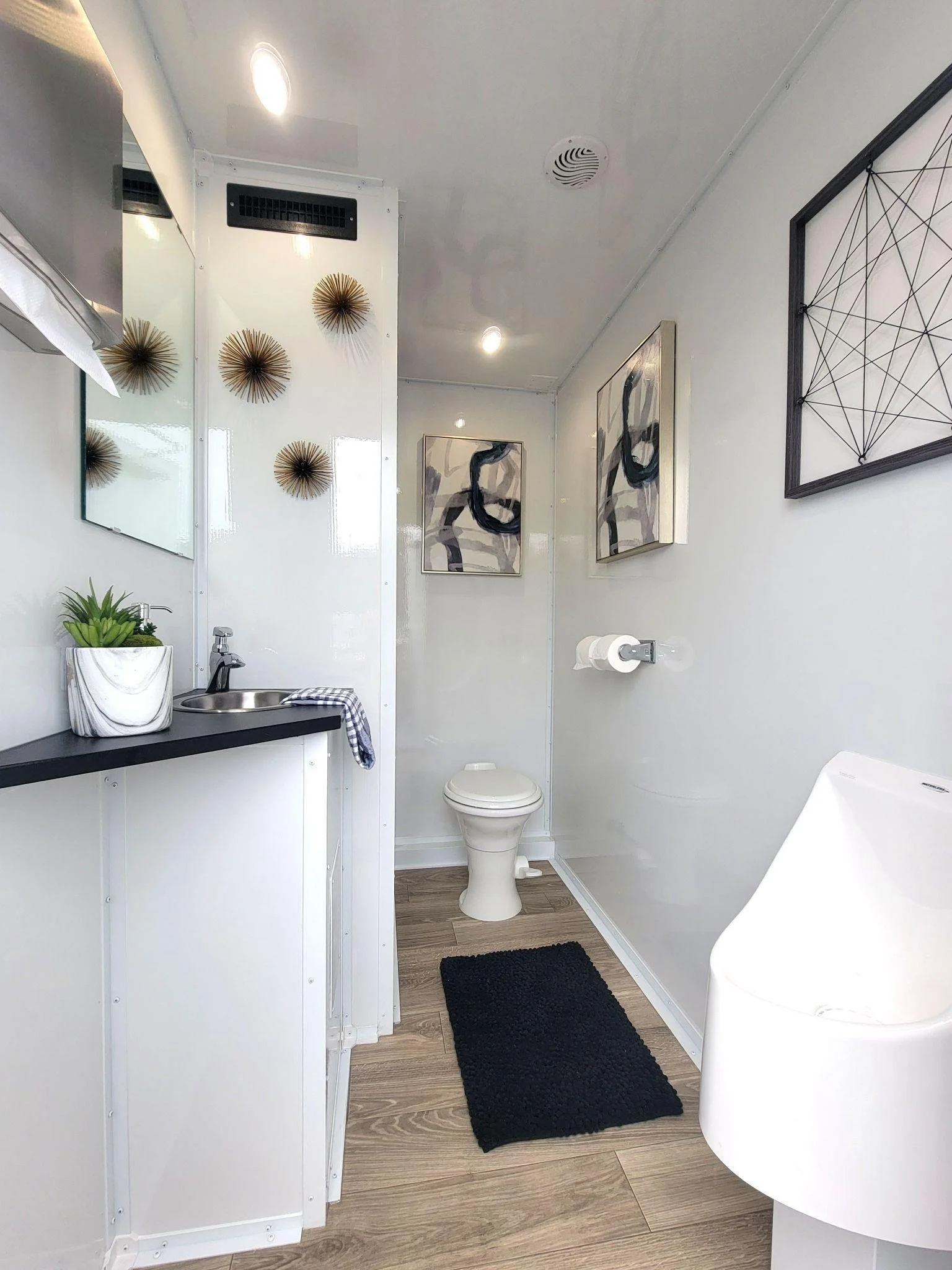 Small, modern bathroom with a black countertop sink, white walls, and decorative wall art. It has a mirror, a black rug, and a white urinal. There are abstract paintings and gold-colored wall decorations.