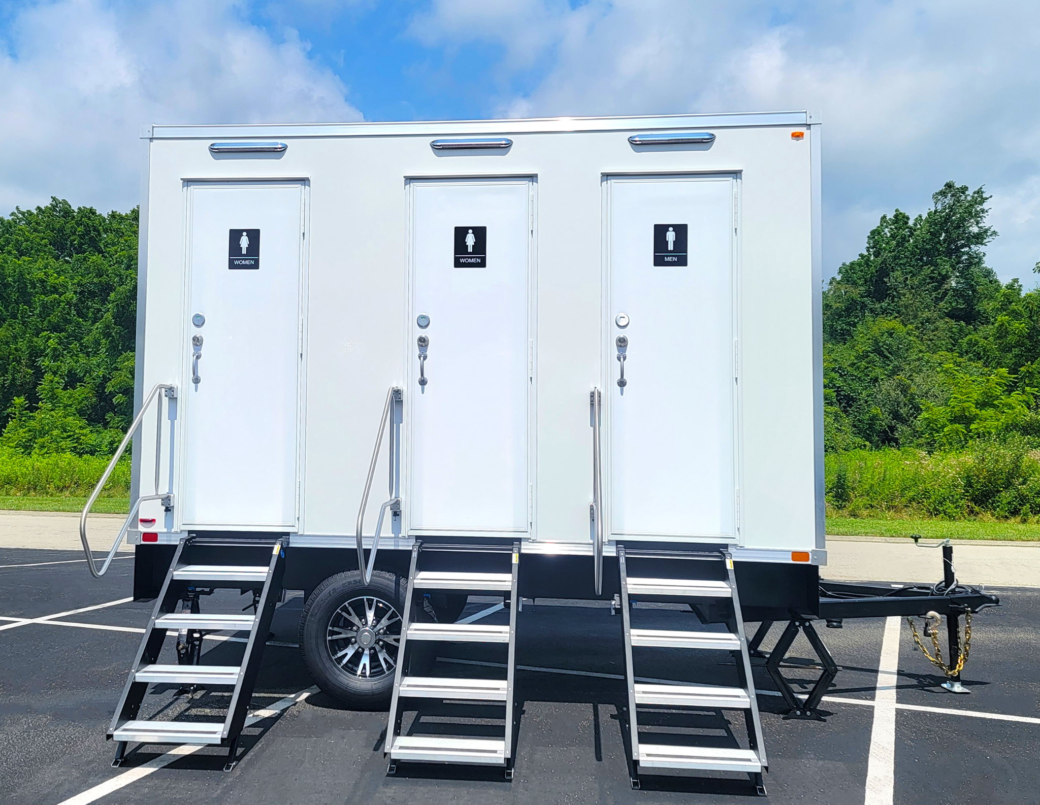 Mobile restroom trailer with separate doors for women's and men's restrooms, three sets of stairs leading up to the doors, parked in a parking lot with trees and a partly cloudy sky in the background.