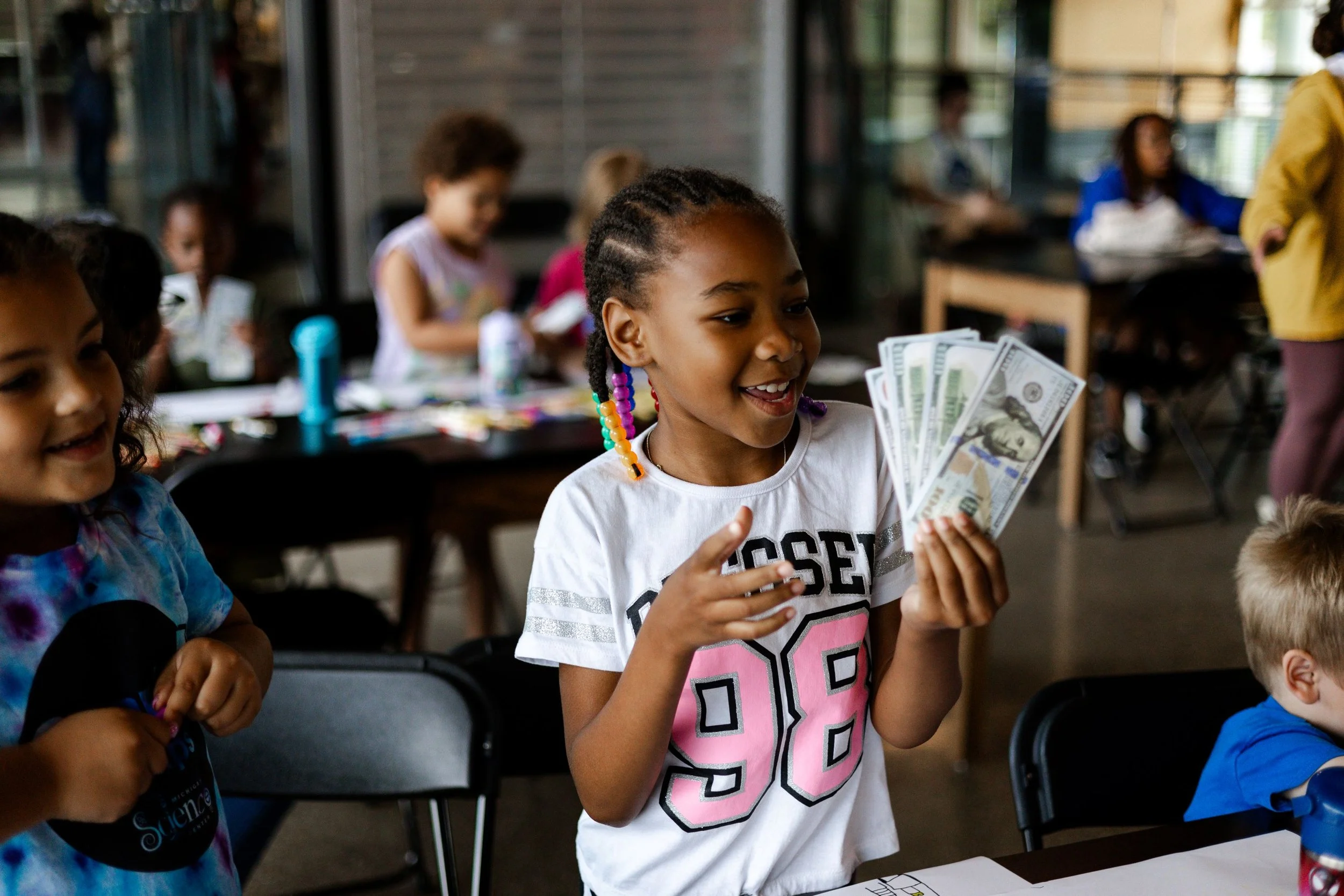 A young girl with rainbow-colored braids holding several hundred-dollar bills, smiling, in a classroom with other children.