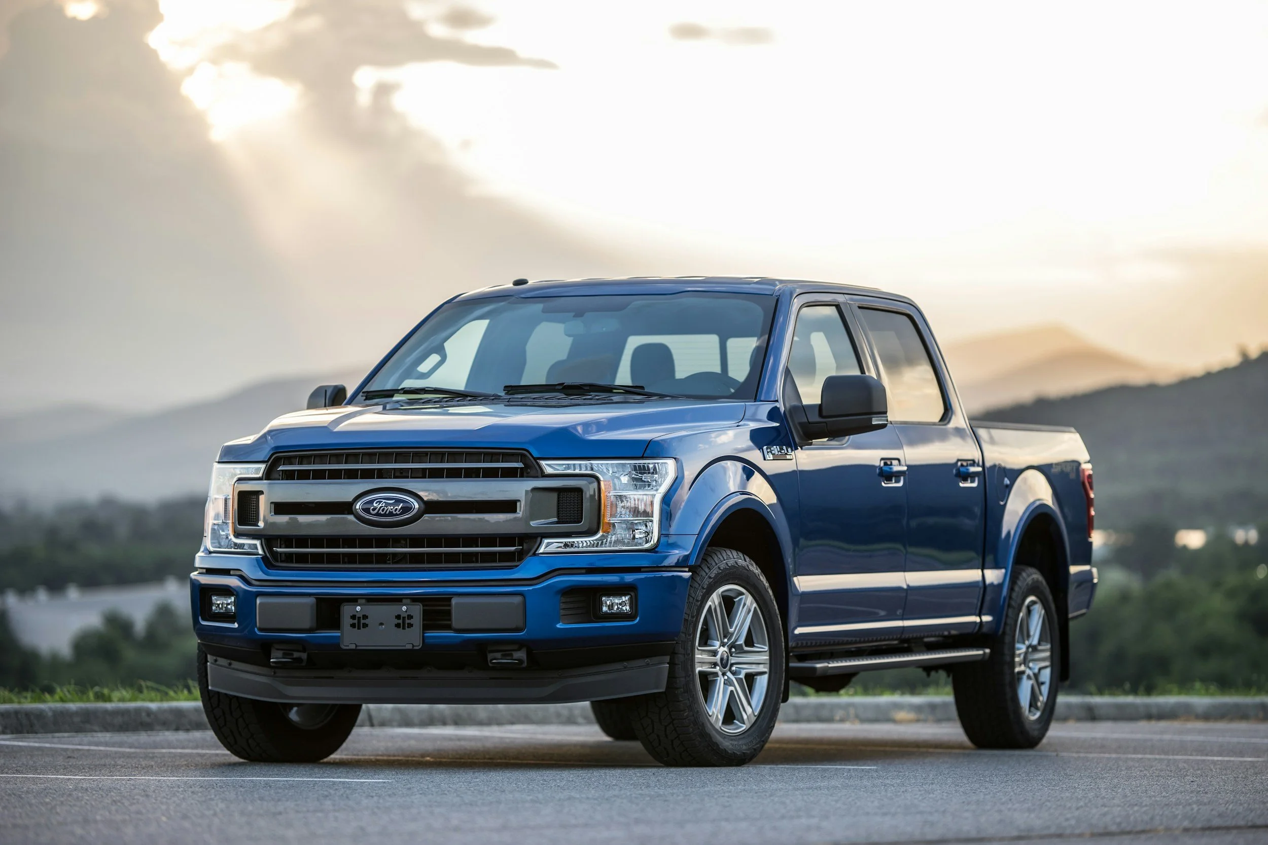 Blue Ford pickup truck parked on a road with a scenic background of mountains and a cloudy sky.