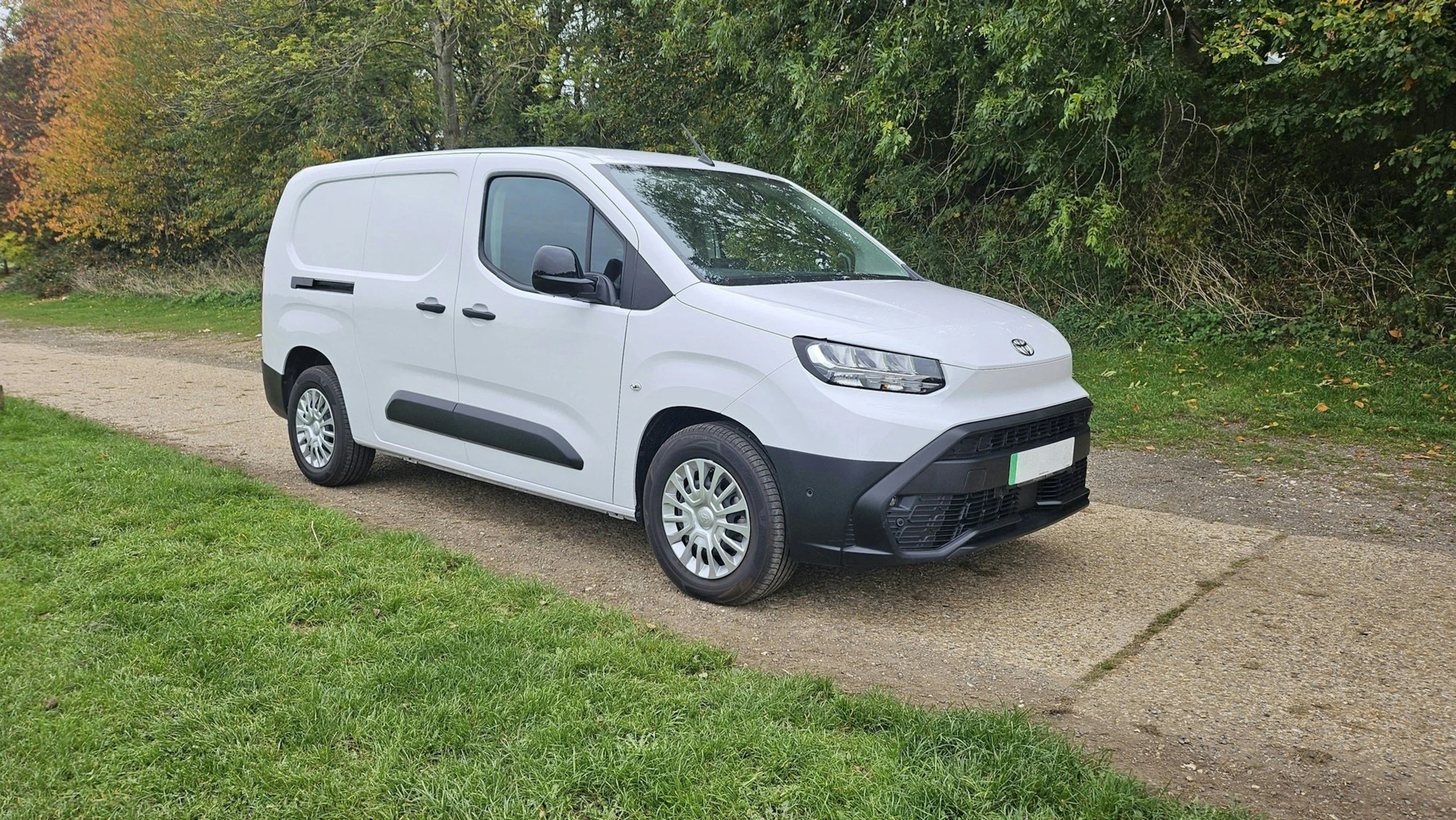 White cargo van parked on a gravel path beside green grass and trees with autumn leaves.