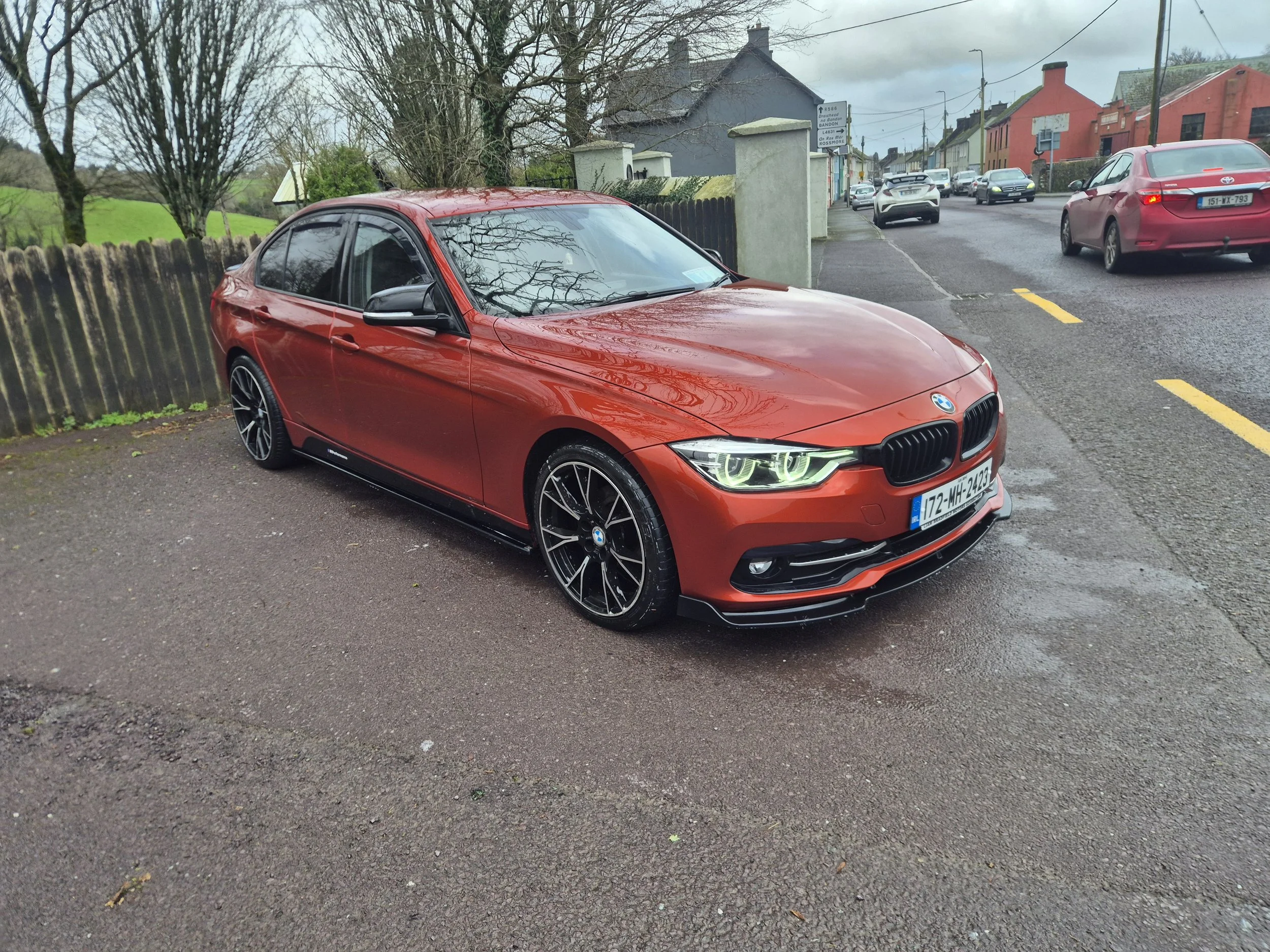 Red BMW parked on a wet roadside in front of a concrete fence with a residential neighborhood in the background, including trees and houses.